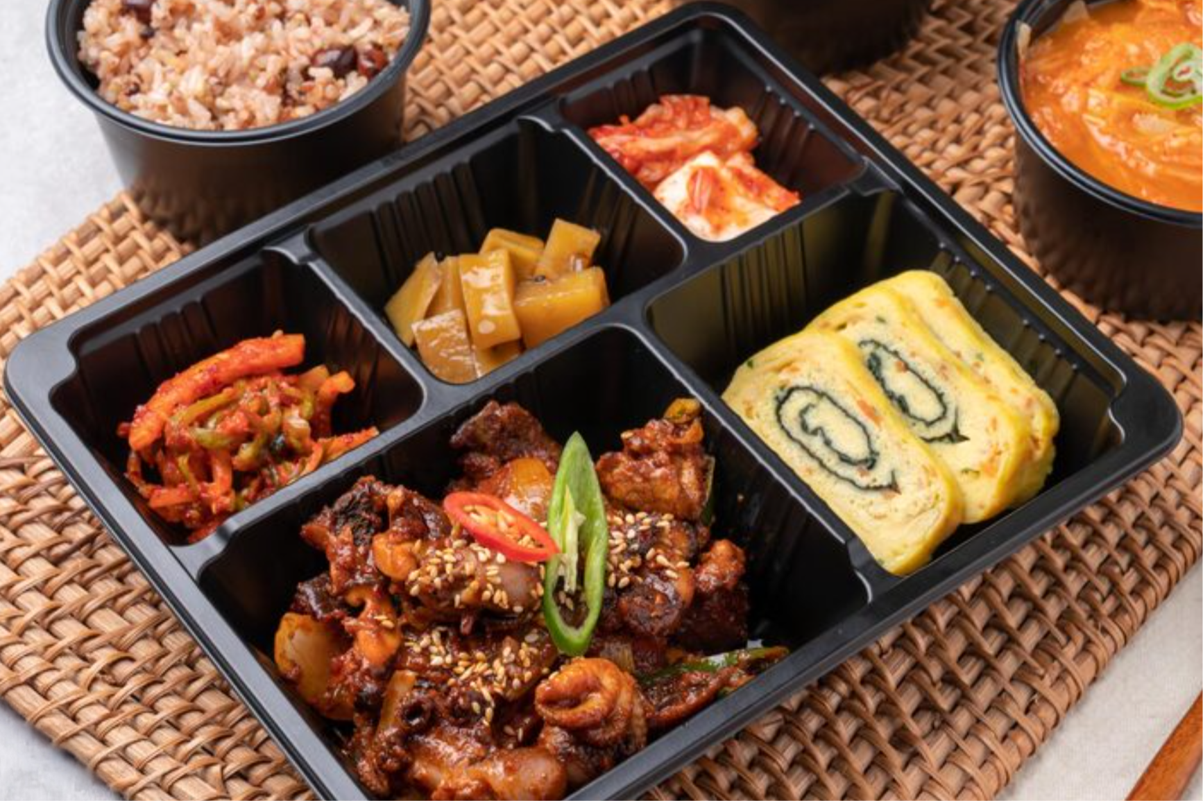 Korean bento box with assorted dishes including spicy chicken, beef bulgogi, rolled omelette, kimchi, pickled vegetables, and side rice, placed on a woven mat.