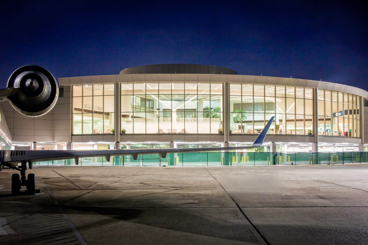 Nighttime view of an airport terminal with large glass windows, illuminated interior, and airplane wing in the foreground.