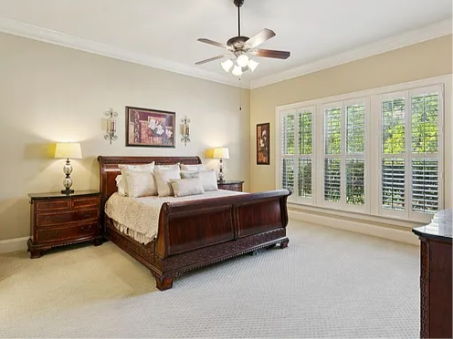 A bedroom with a large wooden bed, matching nightstands, and a dresser. The bed has light-colored bedding and pillows. There are two lamps on the nightstands. A ceiling fan with lights is above. Large windows with white shutters let in natural light.