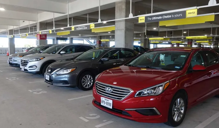 Multiple cars parked in an indoor car dealership lot, with yellow signs hanging from the ceiling displaying the Hertz logo and promotional messages.