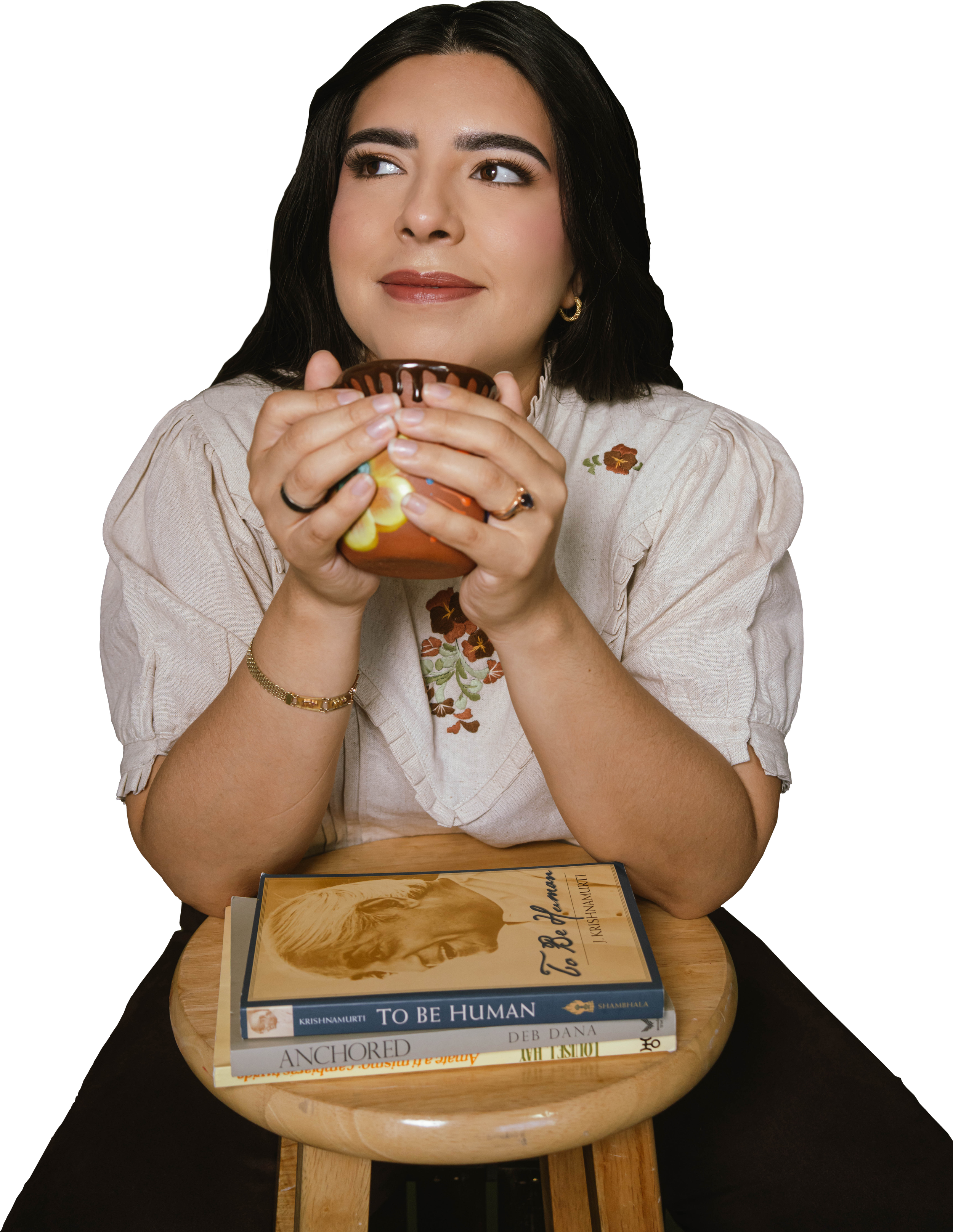 A woman with dark hair and makeup holding a colorful ceramic cup near her mouth, sitting at a round wooden table with a stack of books, including titles like 'To Be Human' and 'Anchored,' in a dimly lit environment.