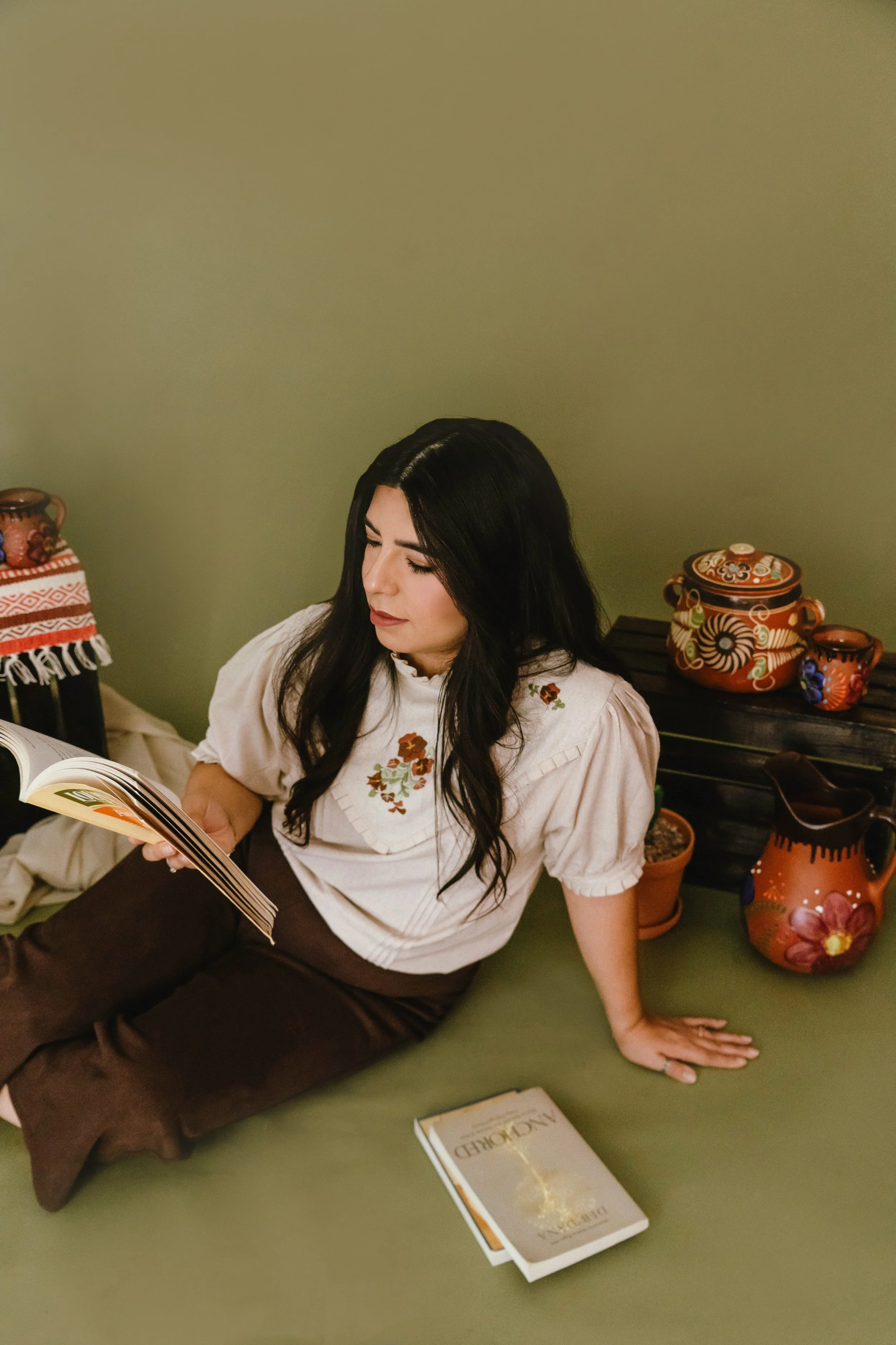 A woman with long dark hair sitting on the floor, wearing a light-colored embroidered blouse and brown pants, reading a book.