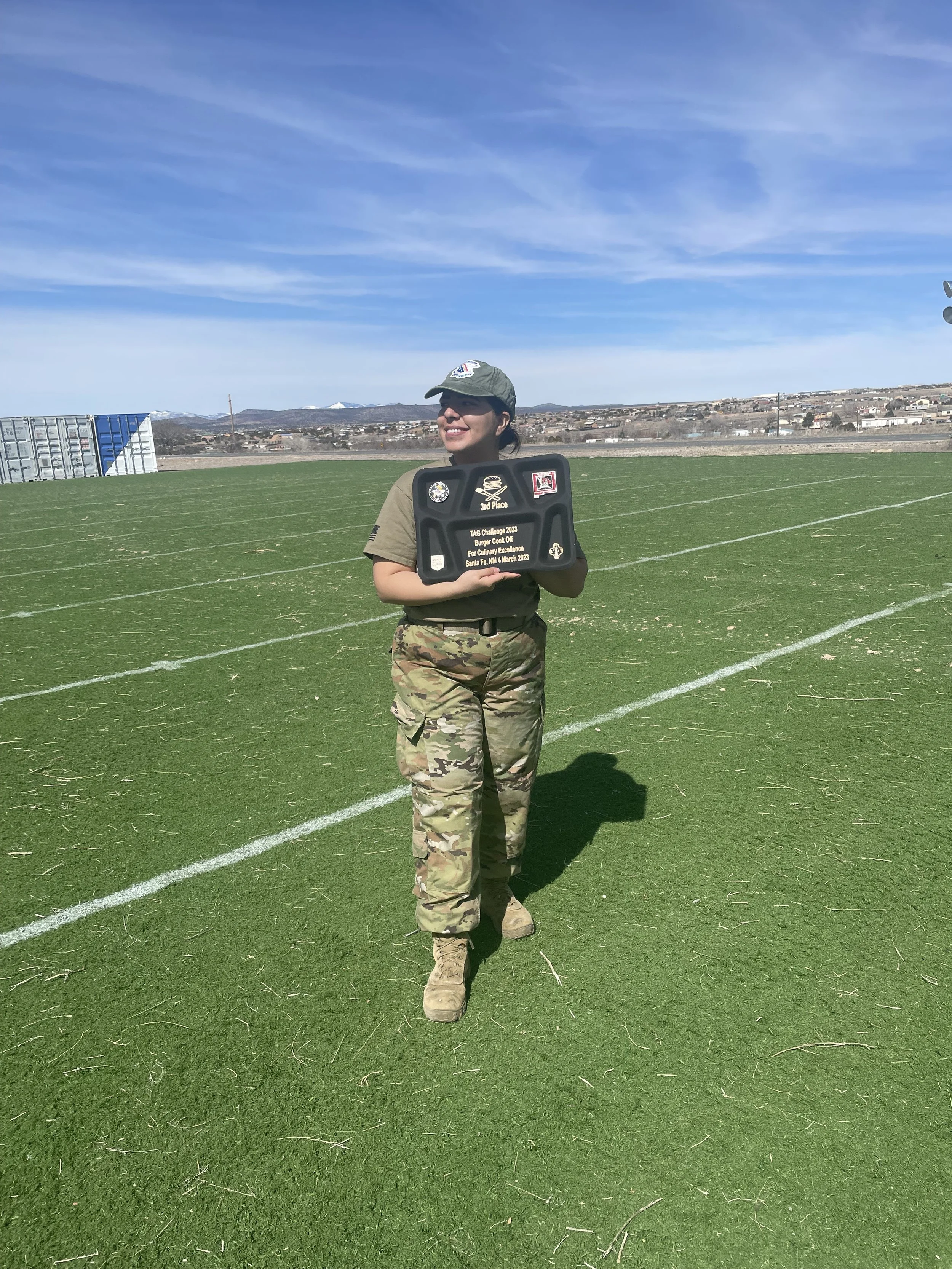 A woman in military camouflage uniform holding a black award plaque on a green sports field with a blue sky, mountains, and a distant cityscape in the background.