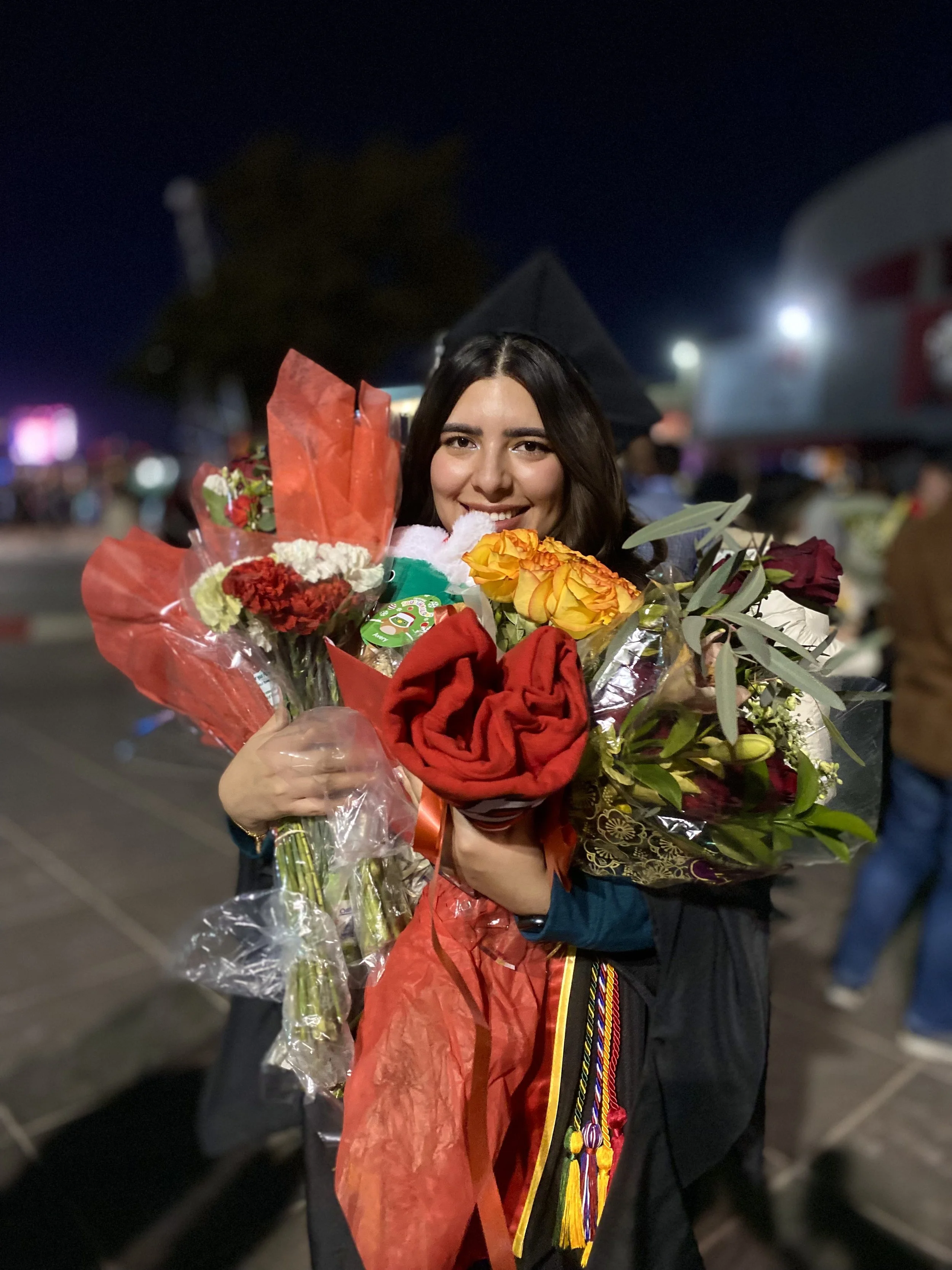 Young woman in graduation cap and gown holding multiple bouquets of flowers during nighttime celebration.