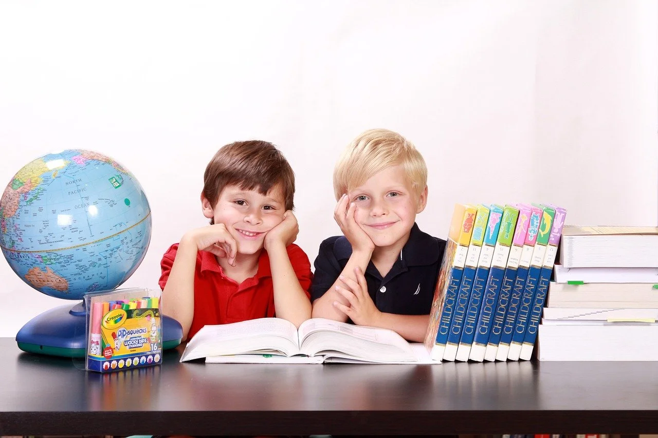 Zwei Jungen sitzen an einem Schreibtisch mit Büchern, Weltkarte, Wasserfarben und einem offenen Buch.