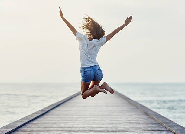 Frau springt auf einem Steg am Strand mit offenen Armen im Sonnenlicht.