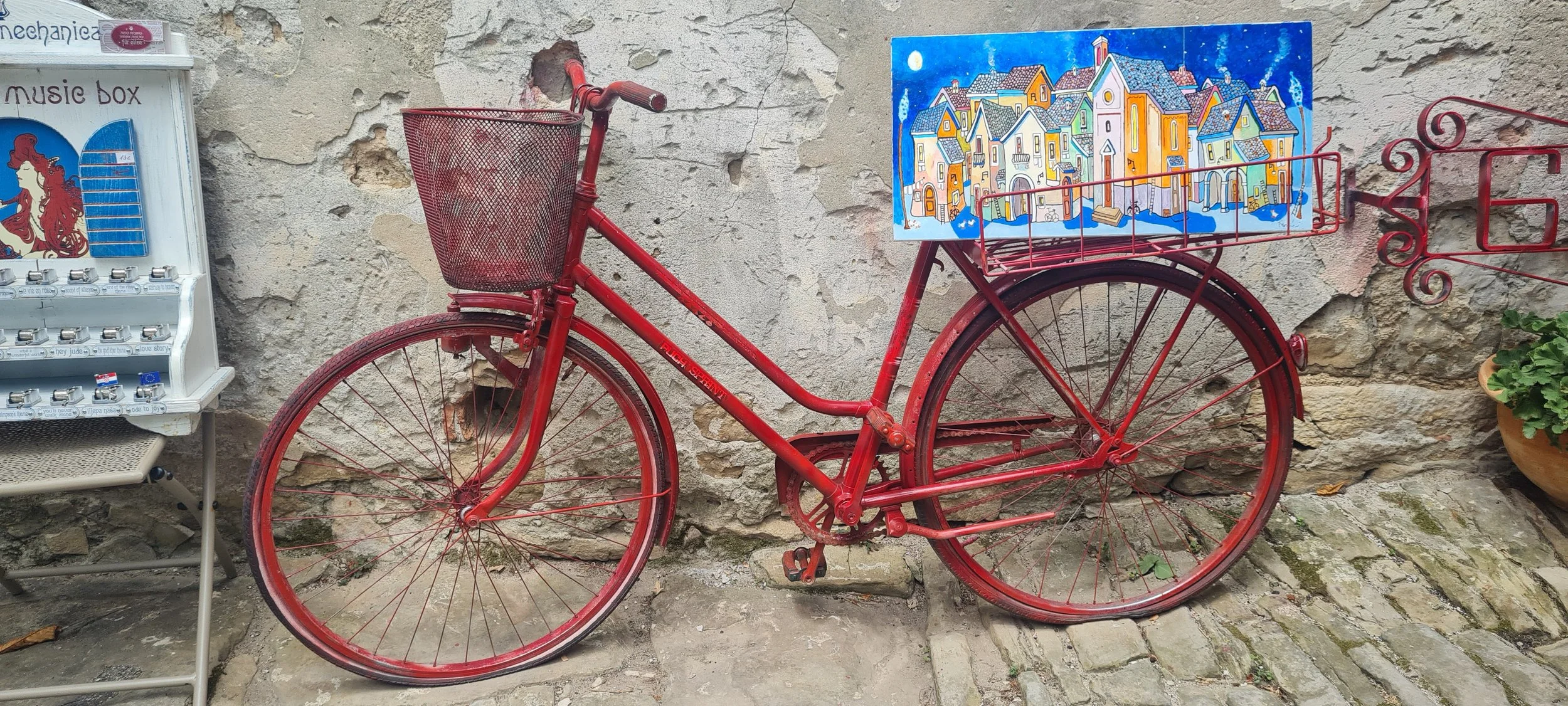 Ein rotes Fahrrad mit einem großen Korb auf einem Kopfsteinpflaster vor einer alten Steinmauer, mit einer bunten Stadtaufnahme als Rahmen im Korb.