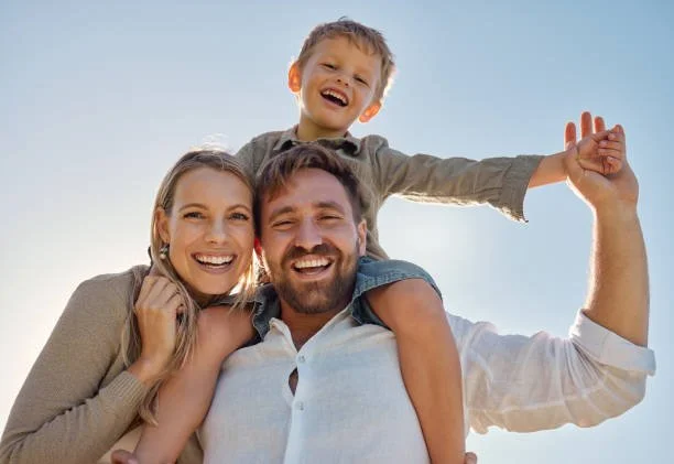 Familie mit einem kleinen Jungen auf den Schultern, alle lachen bei Sonnenschein im Freien.