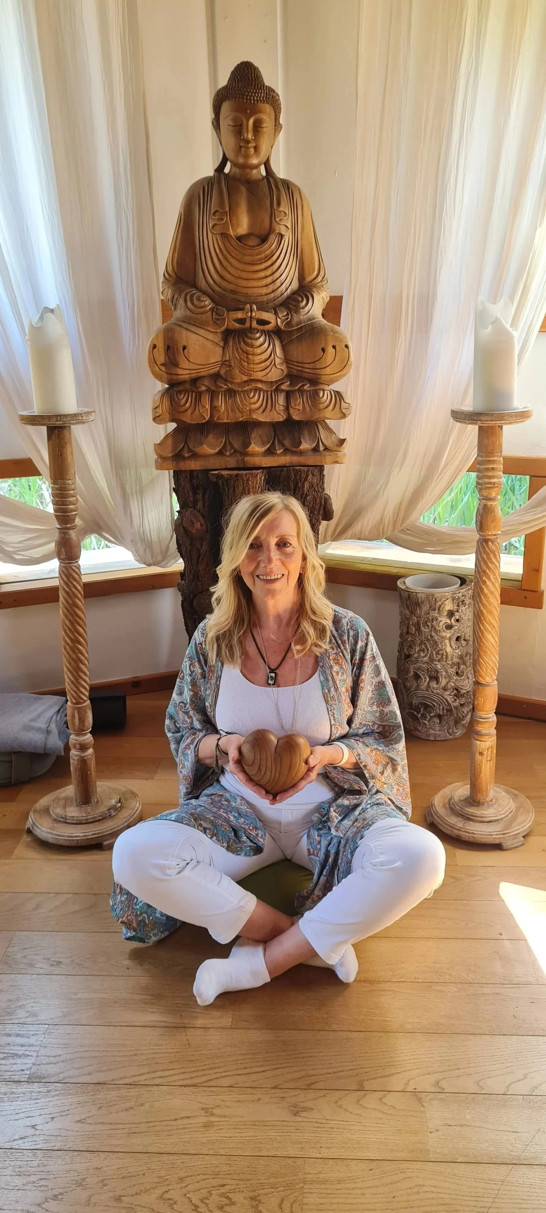 Eine lachende Frau sitzt auf dem Boden, hält ein aus Holz geschnitztes Herz, vor einem großen Holz-Buddha und zwei Kerzenständern.