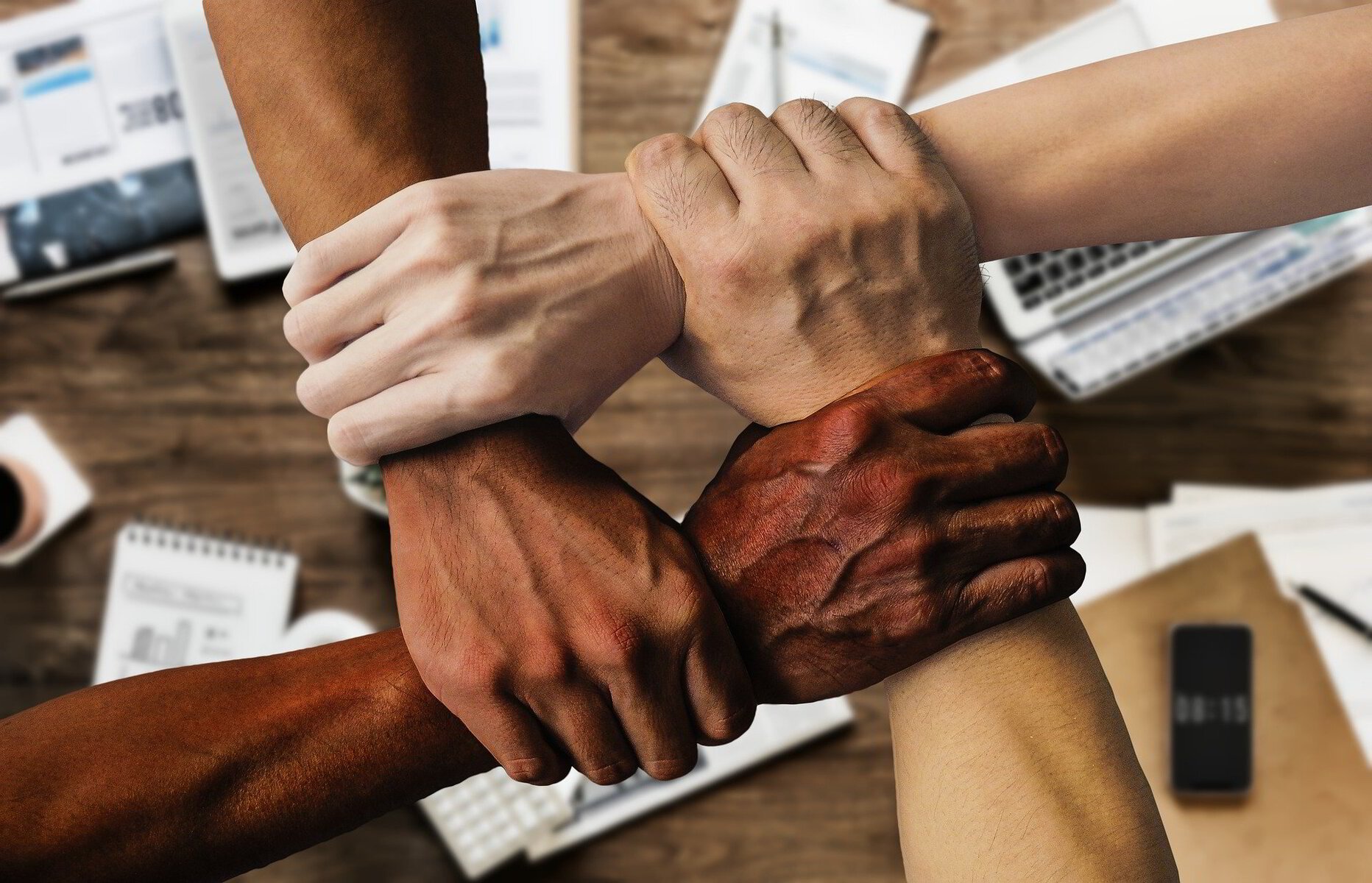 Multiple diverse hands clasped together in a show of unity on a wooden desk surface.