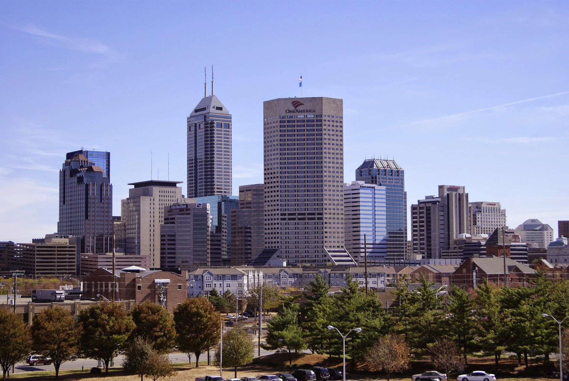 City skyline of Indianapolis with tall skyscrapers, including the OneAmerica building, under a light blue sky with wispy clouds. Foreground features trees, parking lot, and residential houses.