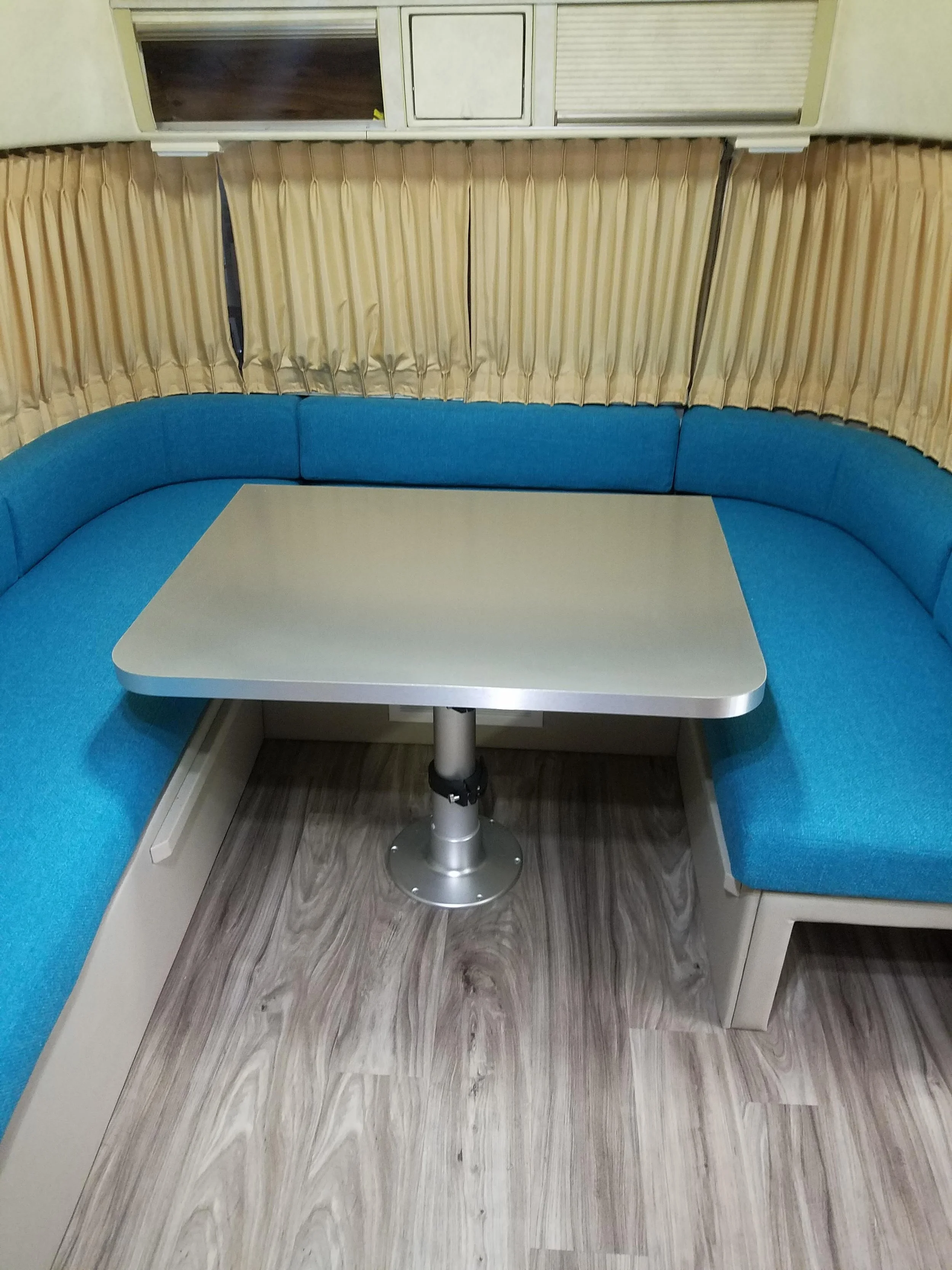 A retro diner booth with curved blue seating, a white table, yellow pleated curtains behind, and a window with a sliding panel above.