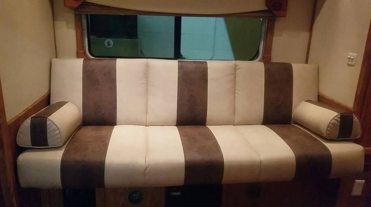 A beige and brown striped sofa with two matching cylindrical pillows is positioned in front of a window within a wooden frame, in a cozy indoor setting.