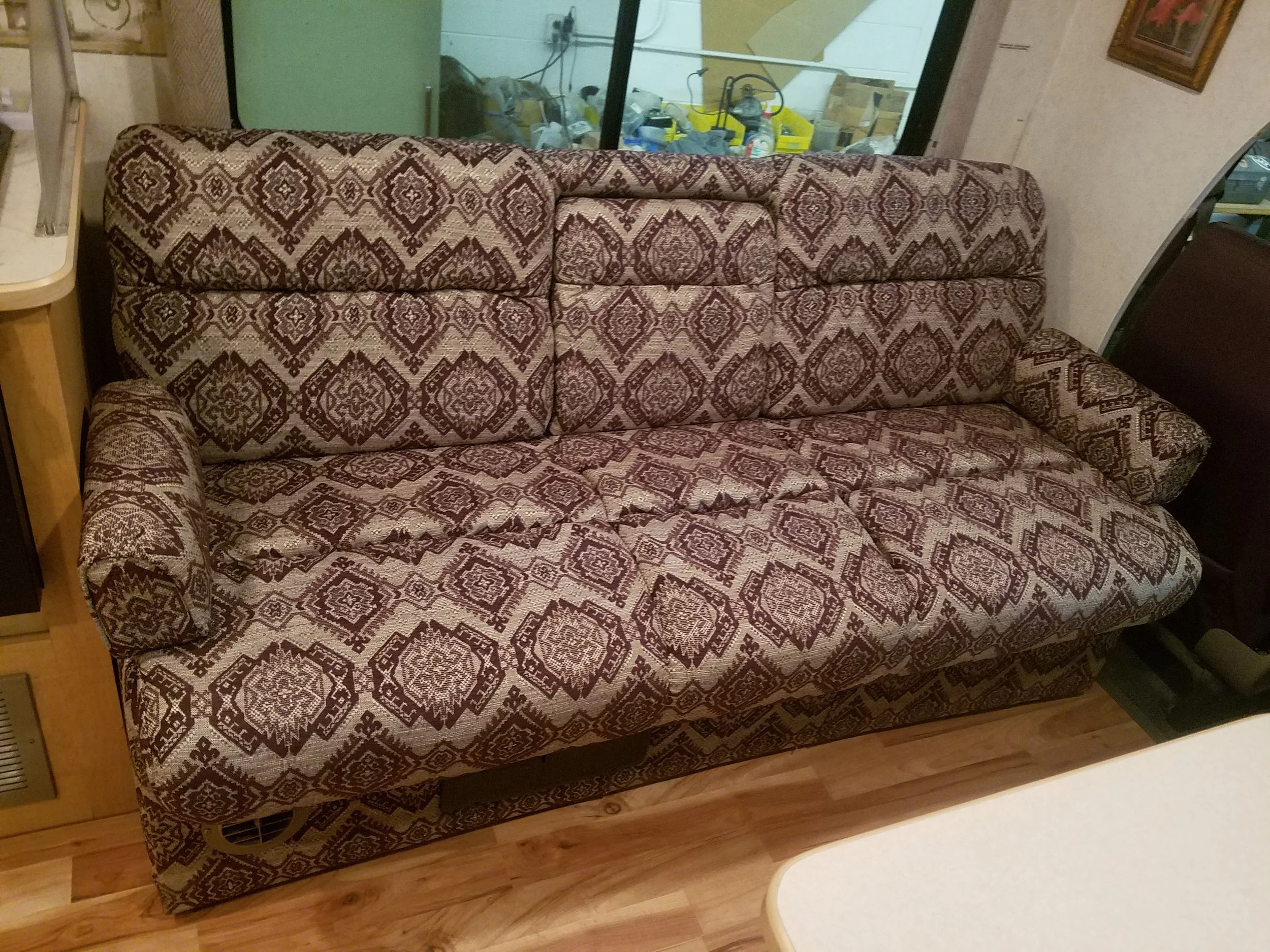 A patterned upholstered sofa with a brown and beige geometric design, placed in a room with wooden flooring and a window showing a storage room with tools and boxes.