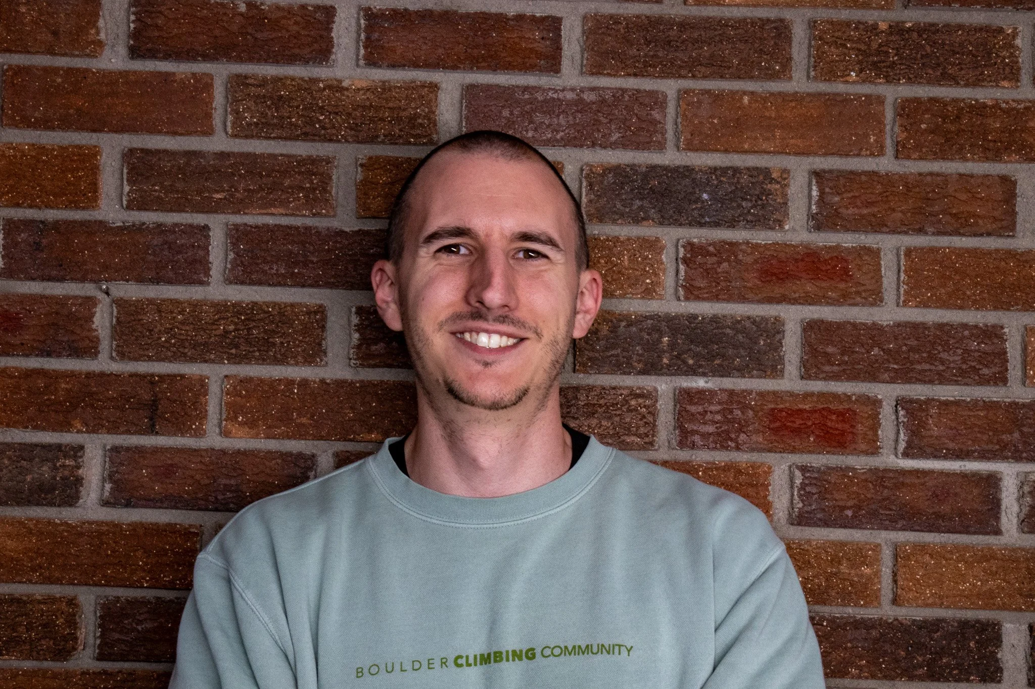 A young man with short hair and a goatee smiling in front of a brick wall, wearing a light gray sweatshirt.