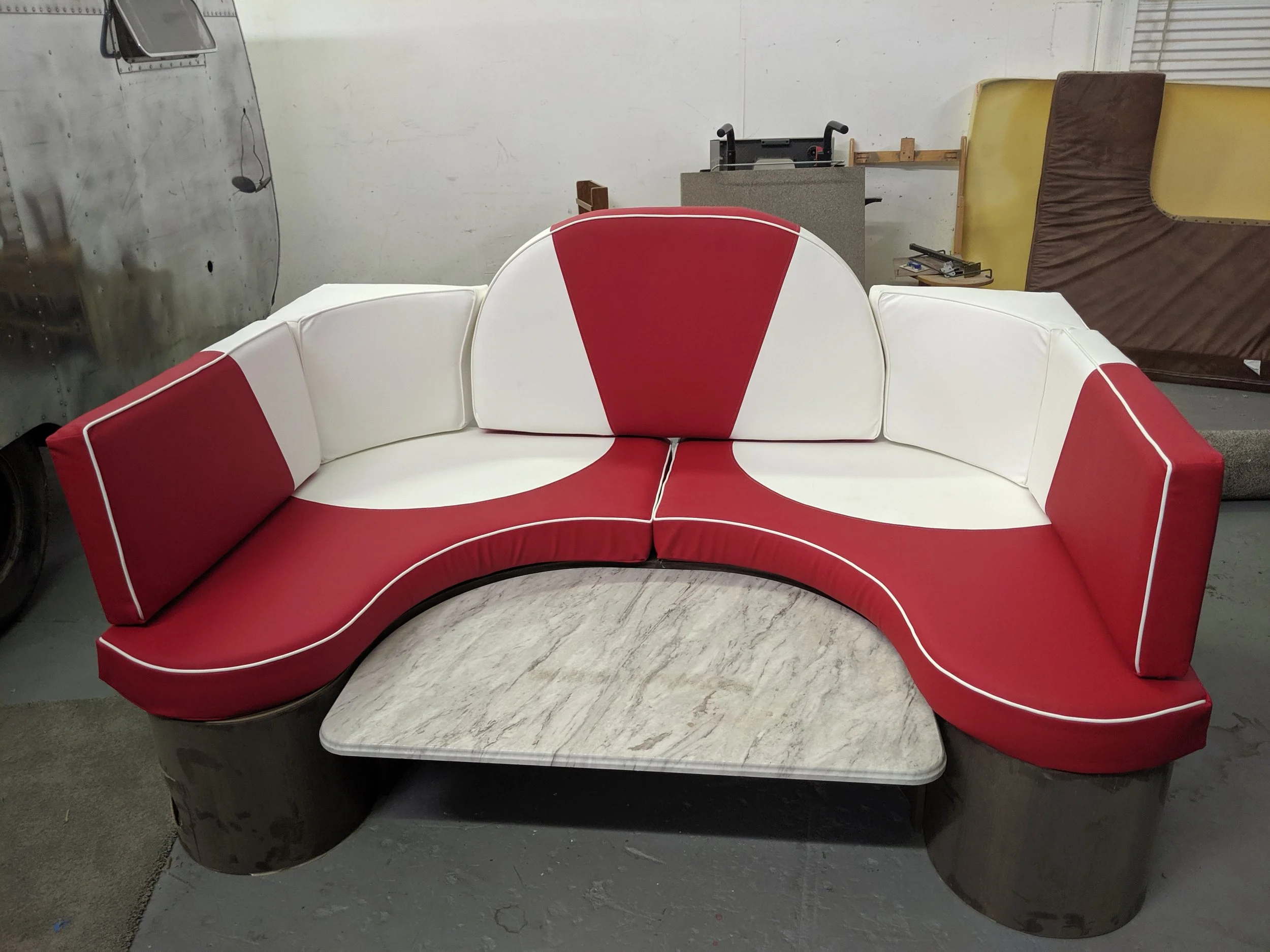 A retro-style red and white curved sofa with a matching white coffee table in front, placed in an industrial-looking space with a vintage aircraft fuselage on the left side and an assortment of chairs and equipment in the background.