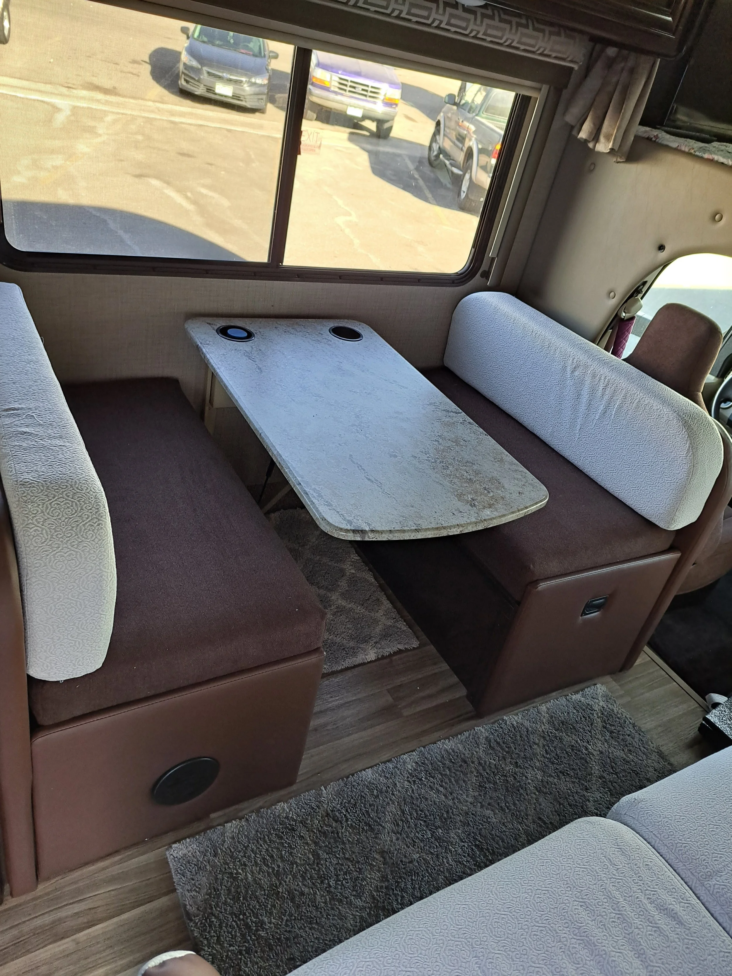 Interior of a recreational vehicle or camper with a small dining area, including a marble-like table, bench seating, and upholstered cushions, with a window showing parked cars outside.