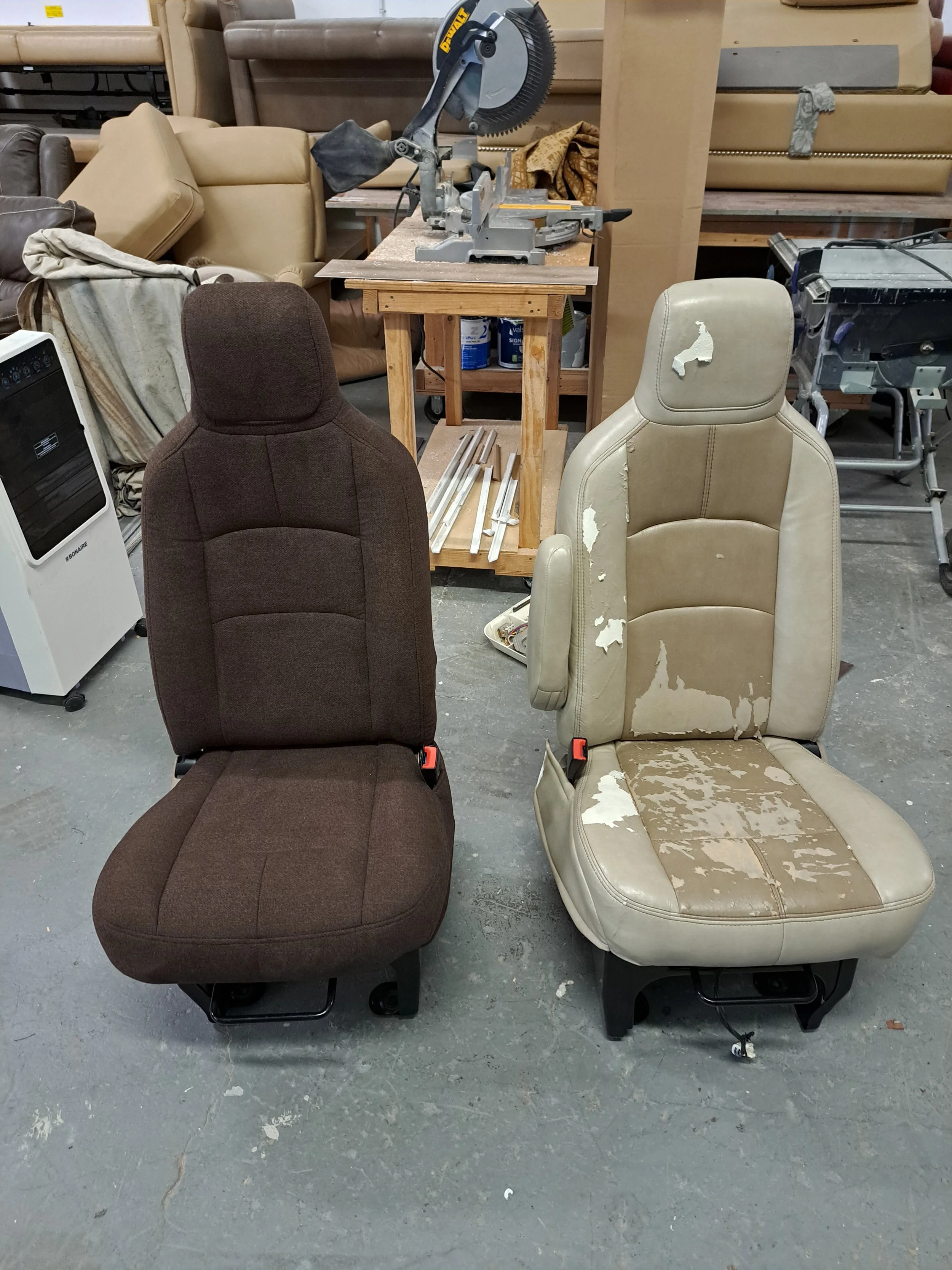 Two car seats, one brown and one beige, are in front of a workshop station. The beige seat is heavily worn and torn, while the brown seat appears in better condition. Behind the seats, there are various furniture pieces, a saw on a stand, and other workshop tools and supplies.