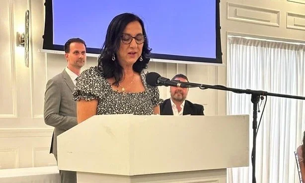 A woman is speaking at a podium with a microphone, three men standing behind her, and a large screen above them in a formal indoor setting.