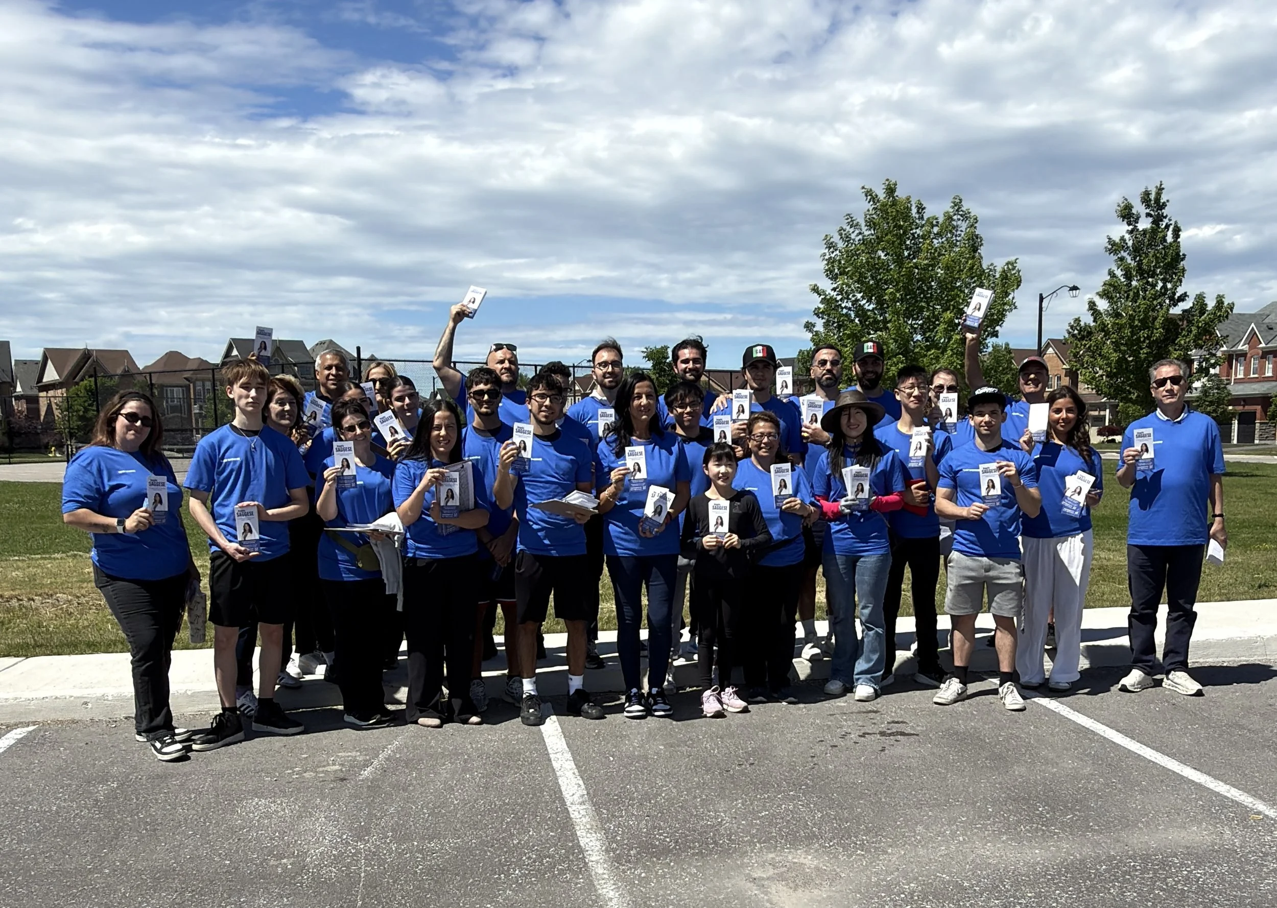 A large group of people standing outdoors in a parking lot, holding up pamphlets or booklets, wearing blue shirts, with houses and a tree in the background under a partly cloudy sky.