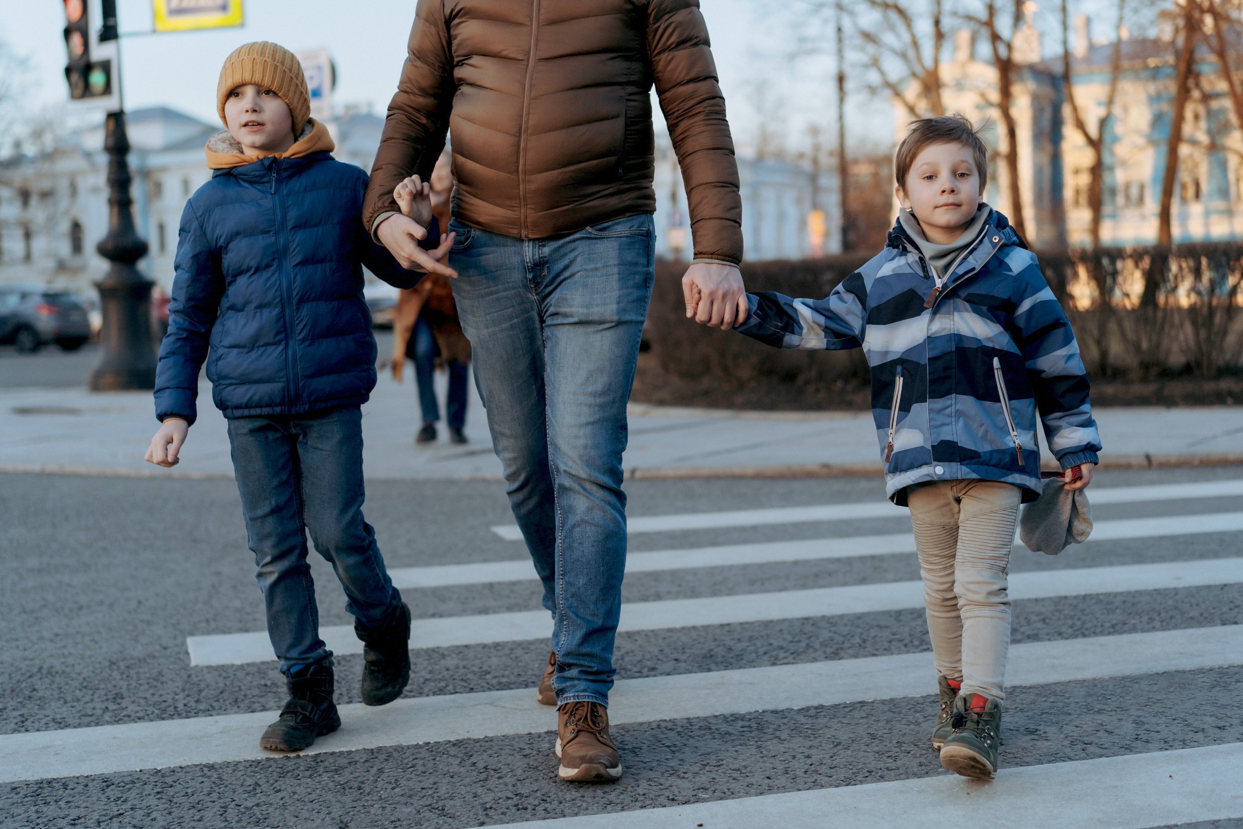 Two young boys and an adult woman crossing a street at a crosswalk in an urban area. The woman is holding both boys' hands. The boys are dressed in jackets and boots, and the woman is wearing a brown puffer jacket and jeans. Trees with autumn leaves and buildings are visible in the background.