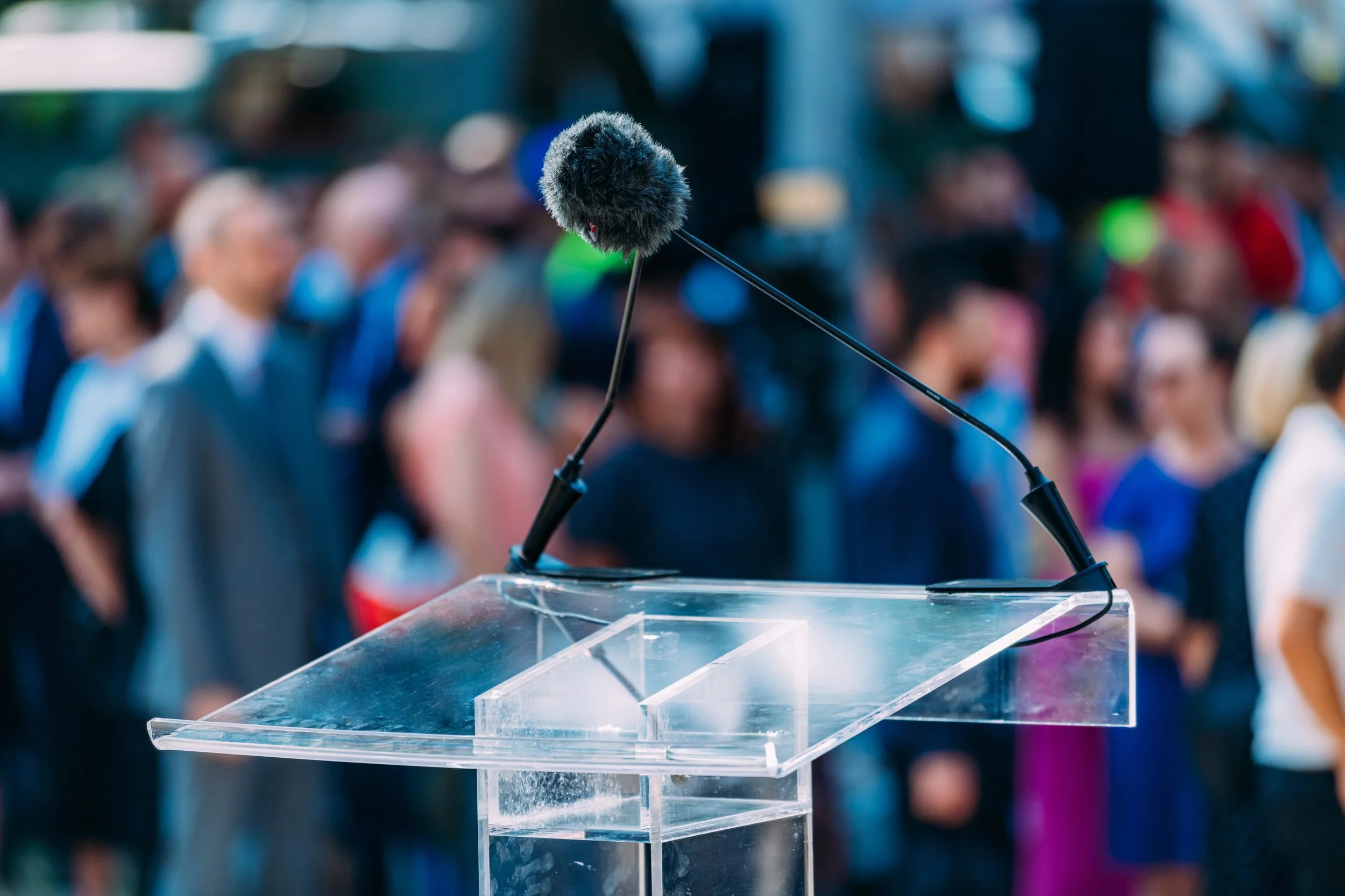 Fuzzy microphone on a stand at a podium with a crowd of blurry people in the background.