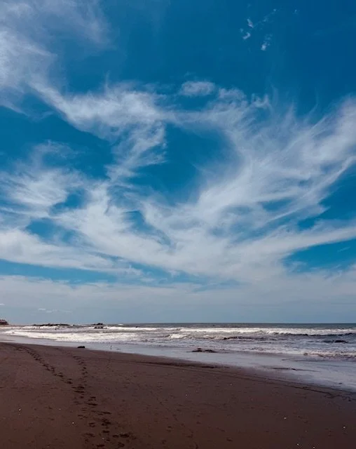 Empty beach with footprints in the sand, ocean waves, and a blue sky with wispy clouds.