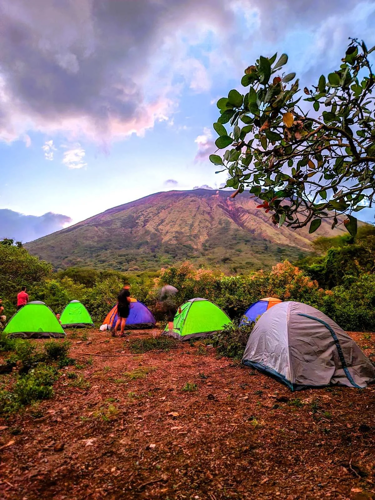 Camping scene with multiple brightly colored tents on a dirt area in front of a mountain. People are gathered around, with some standing and others near the tents. San Cristobal volcano in the background with a partly cloudy sky overhead.