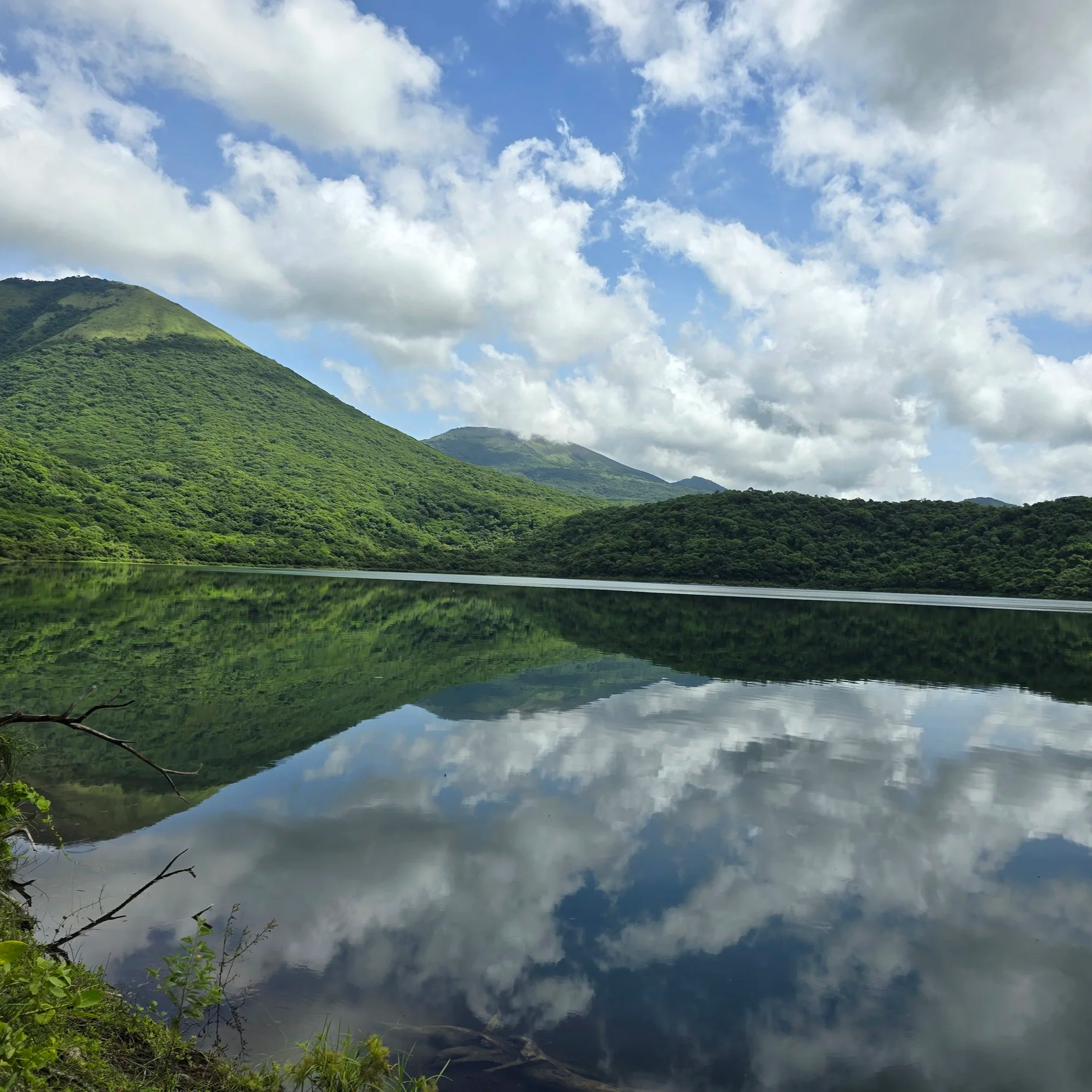 Mountains covered in green trees reflected in a calm lake under a partly cloudy sky.
