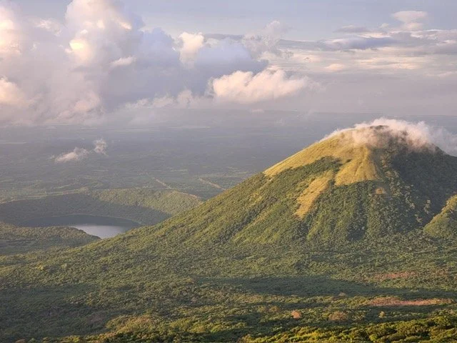 A lush green volcano with a slight plume of smoke at the top, surrounded by forested landscape and cloudy sky.