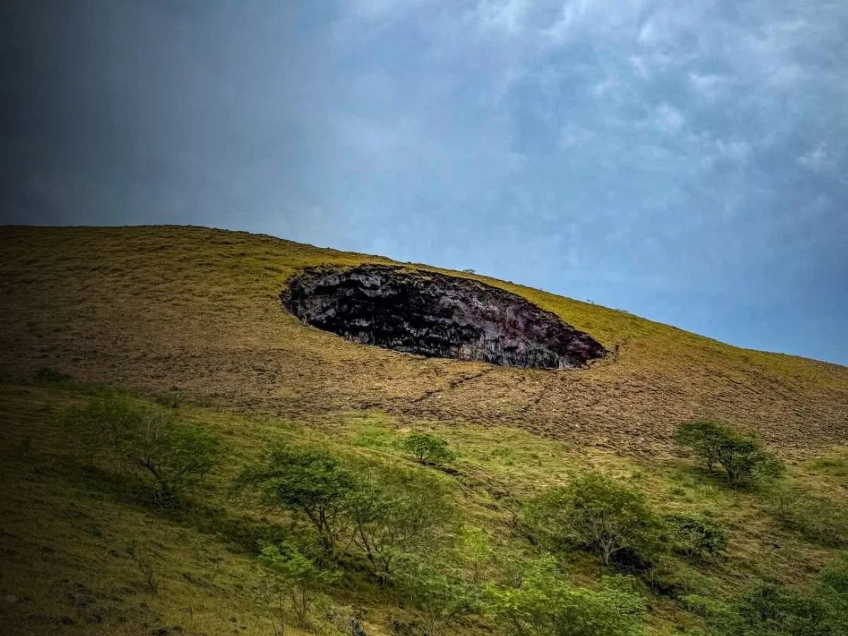 A hillside with a large, sinkhole on its slope, partially surrounded by grass and a few small trees, under a blue sky with some clouds.