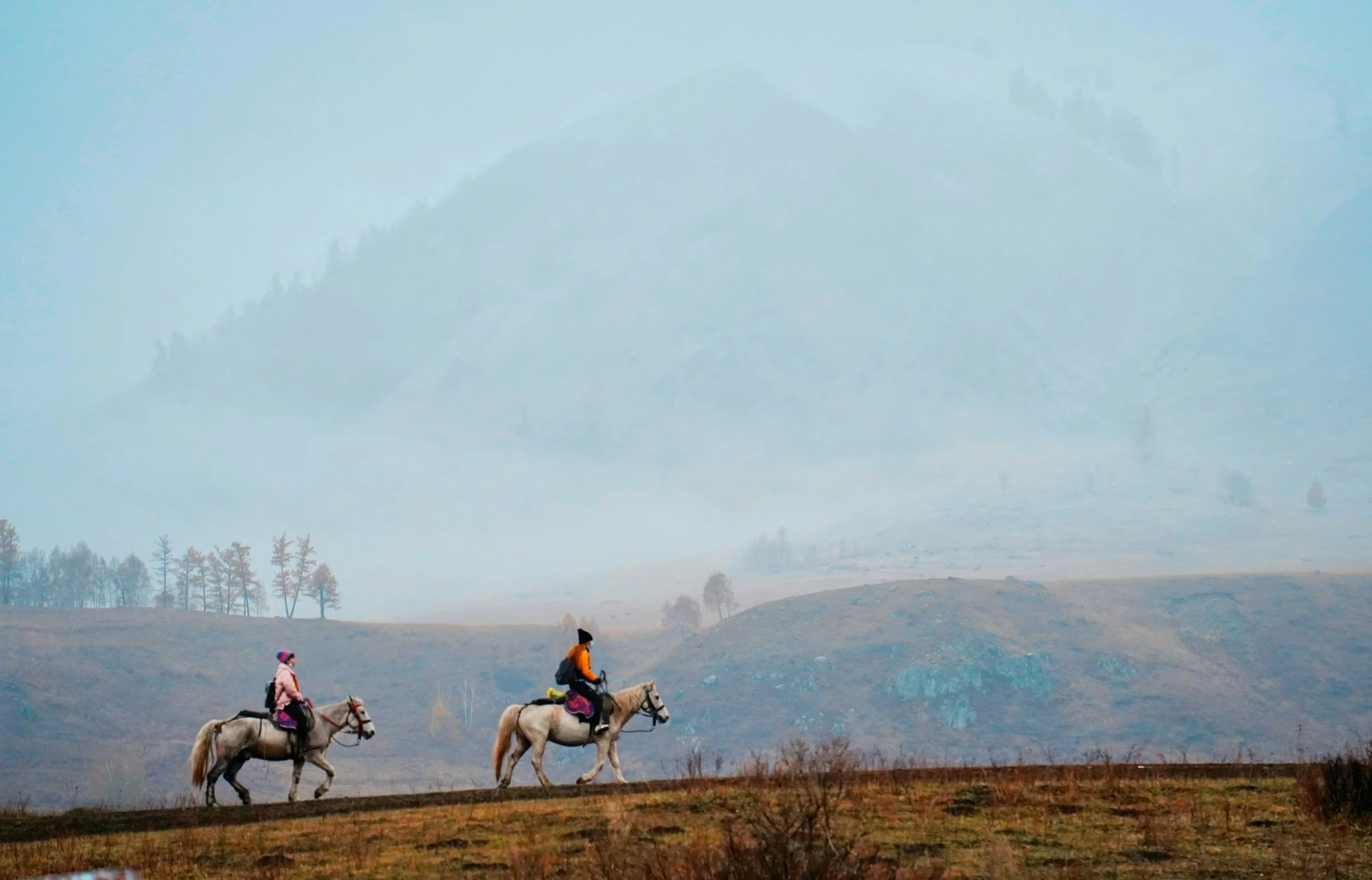 Two children riding horses in a rural landscape with a mountain in the background, some trees, and a cloudy sky.