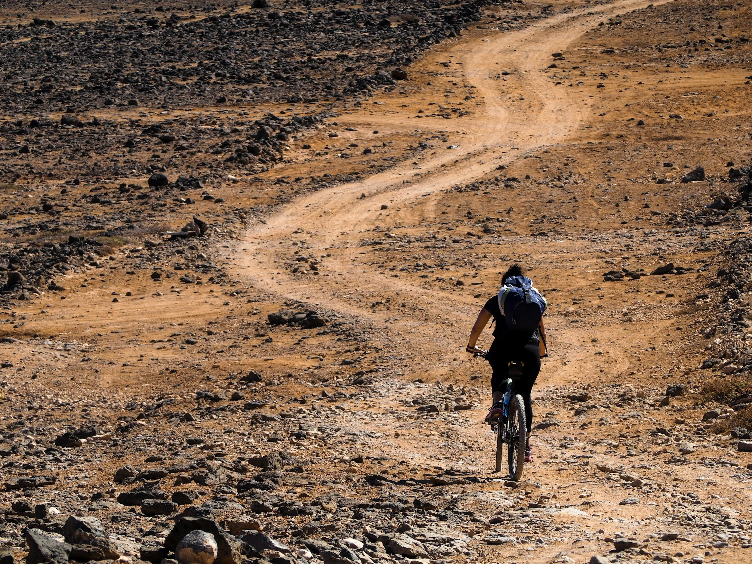 A person riding a mountain bike on a dirt trail through a rugged, rocky, desert landscape with barren hills in the background.