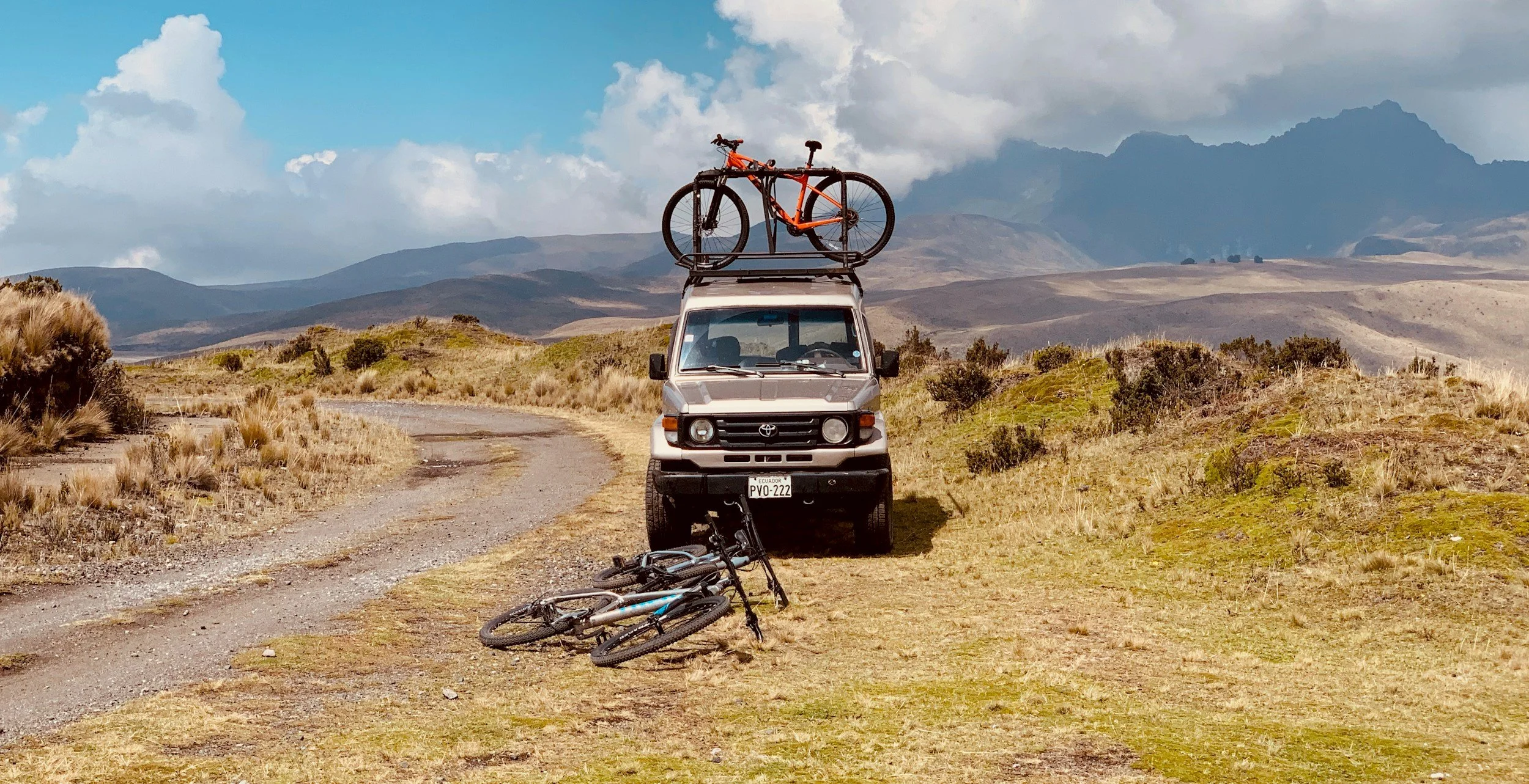 A white van parked on a dirt trail in a mountain landscape, with two bicycles on the ground in front and a bike mounted on the roof. Mountains and clouds in the background.