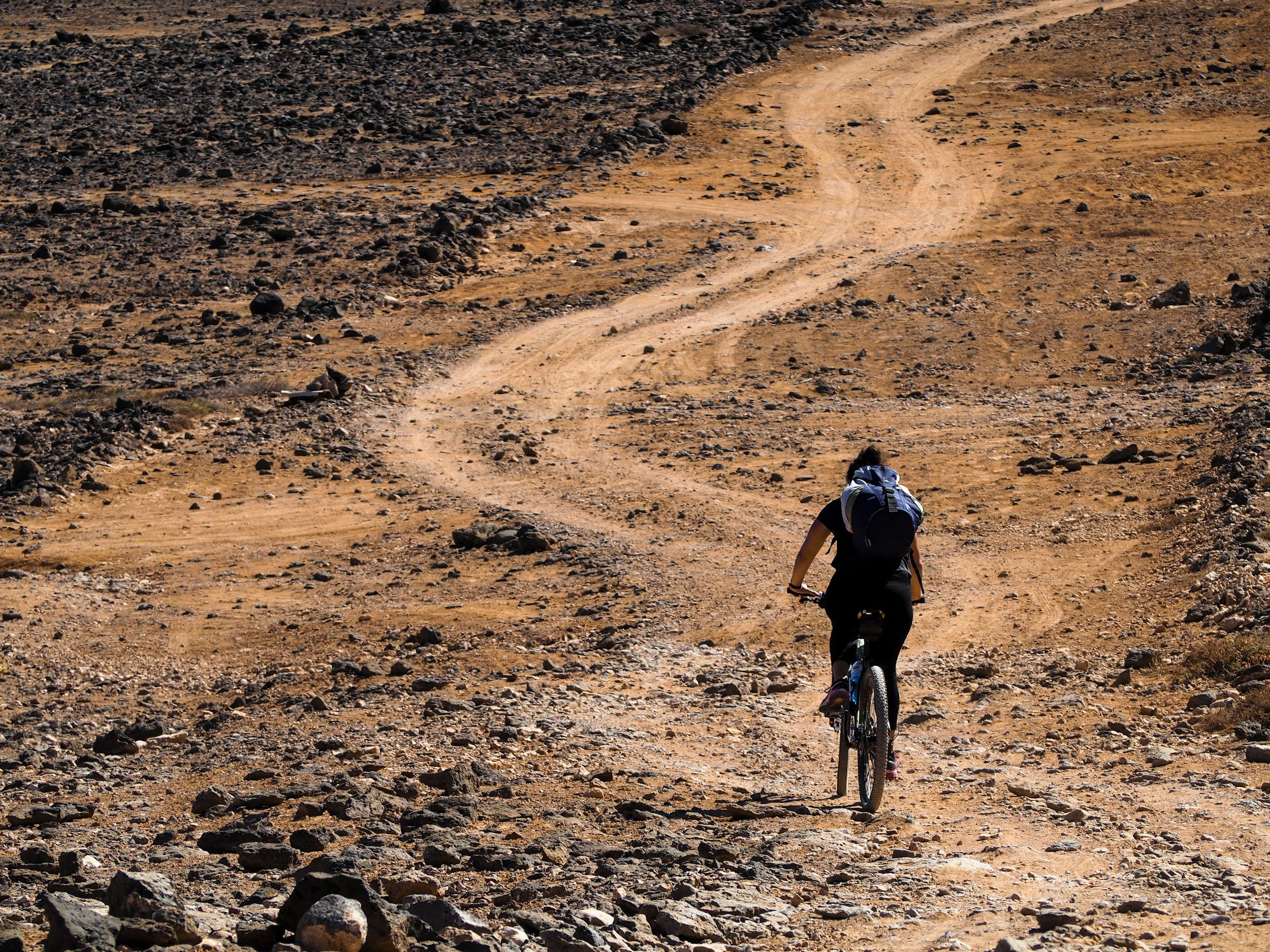 A person riding a mountain bike on a rugged dirt trail in a dry, rocky landscape.