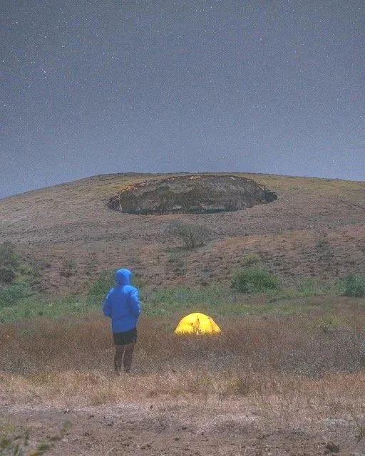 Person in a blue jacket stands near a yellow tent on a grassy field at night, looking at a volcano under a starry sky.