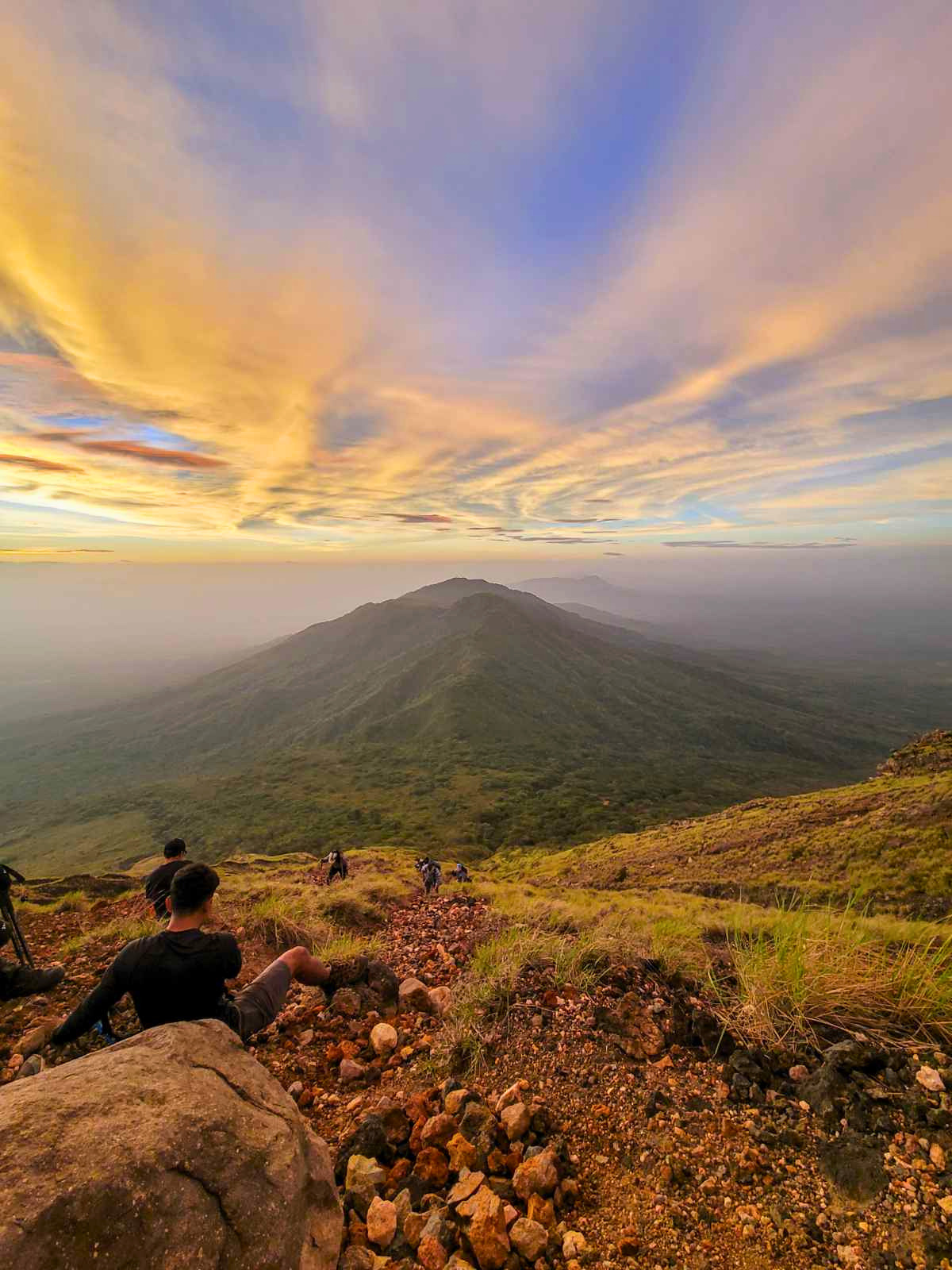 A group of hikers resting on the rocky and grassy hillside of a volcano during sunset, with a view of distant volcanoes, lush green terrain, and a cloudy sky with warm orange and purple hues.