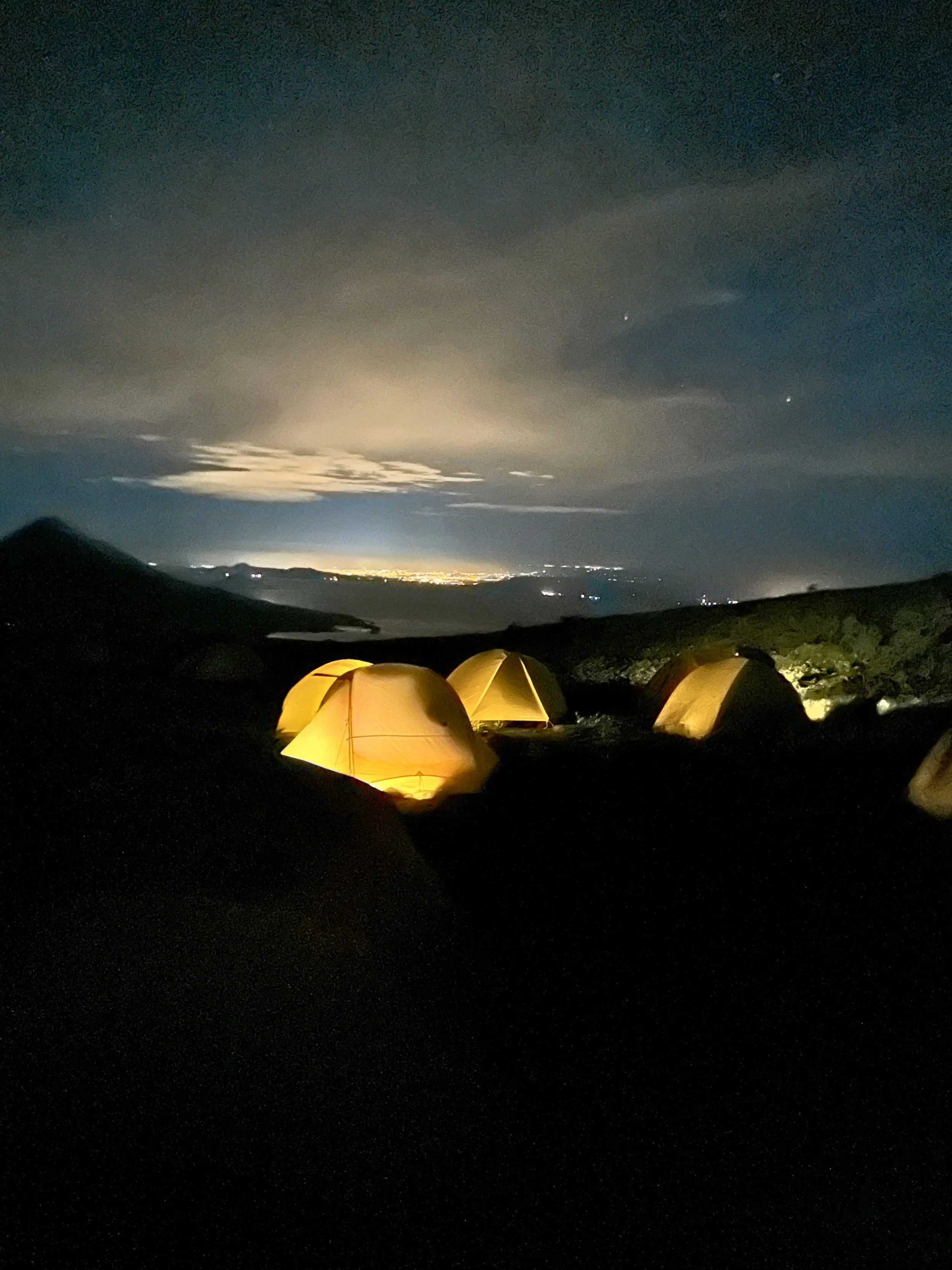 Nighttime landscape with illuminated tents in the foreground, volcanoes in the distance, and a bright glow in the sky near the horizon, possibly from moonlight or city lights.