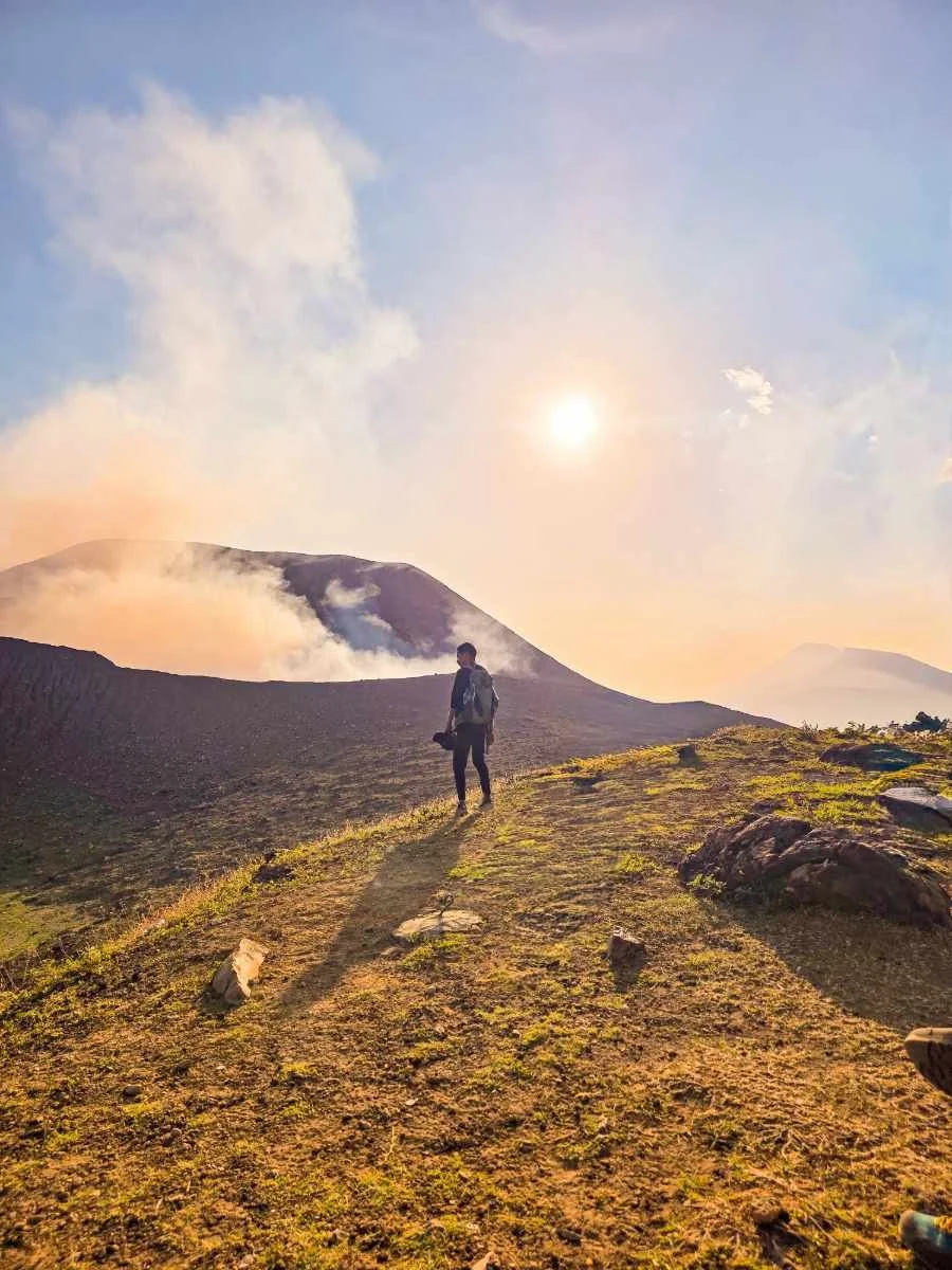 A hiker walking on a rocky trail in a volcanic landscape during sunset, with clouds and smoke emanating from the volcano in the background.