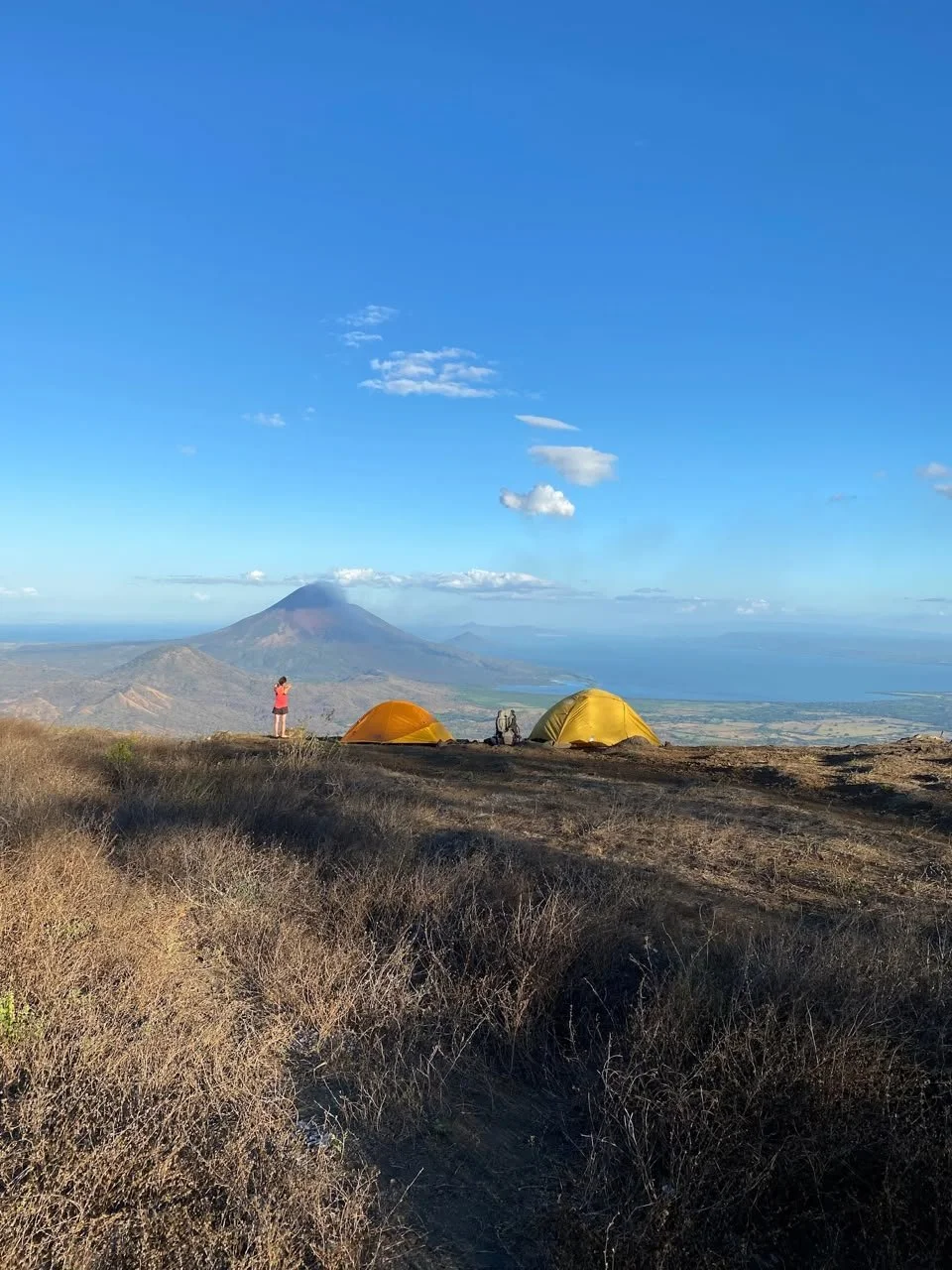 Two tents, one yellow and one orange, set up on a mountain overlooking a valley with a person taking a photo. In the background is a prominent volcano, with a blue sky and scattered clouds.