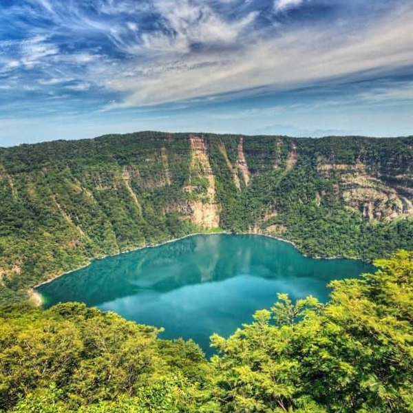A scenic view of cosiguina turquoise crater volcano lake surrounded by lush green forested hills and steep cliffs under a partly cloudy sky.