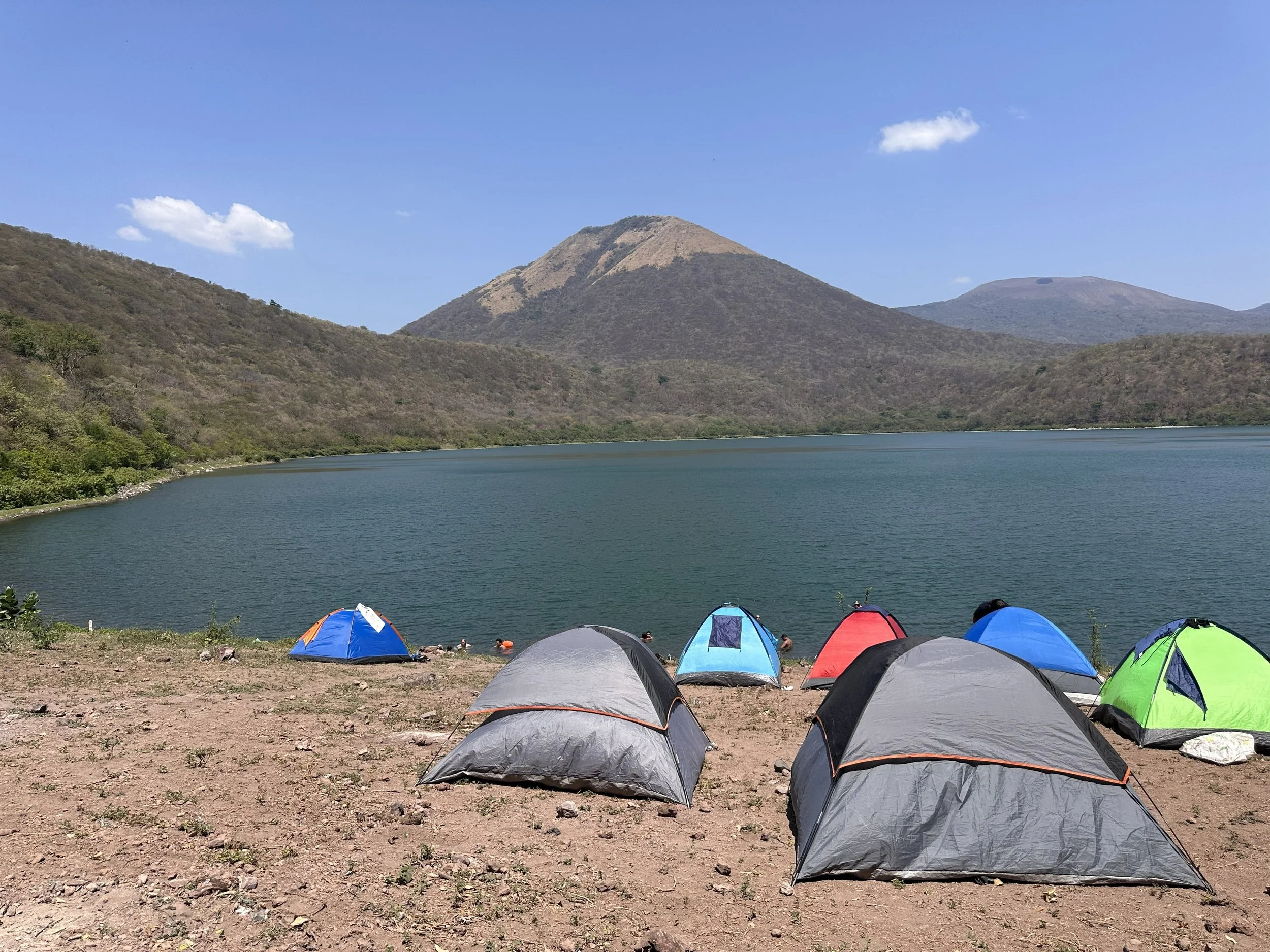 A lakeside campsite with colorful tents on the shore of a body of water, surrounded by mountains and a blue sky with a few clouds.