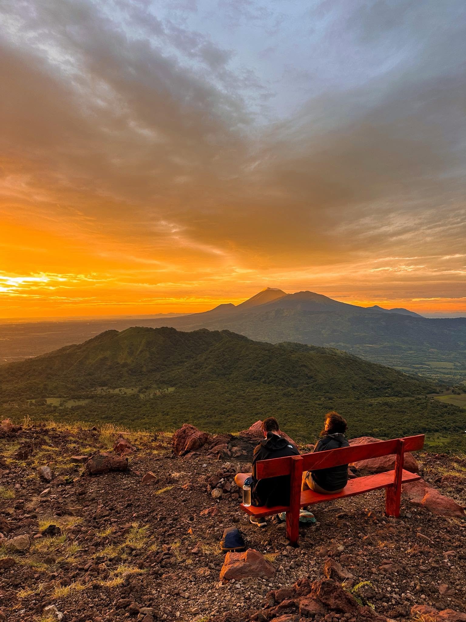 Two people sit on a red bench on Telica volcano veiwing the landscape at sunset. The sky is filled with clouds illuminated by orange and purple hues, with a prominent volcano in the background and a smaller peak nearby.
