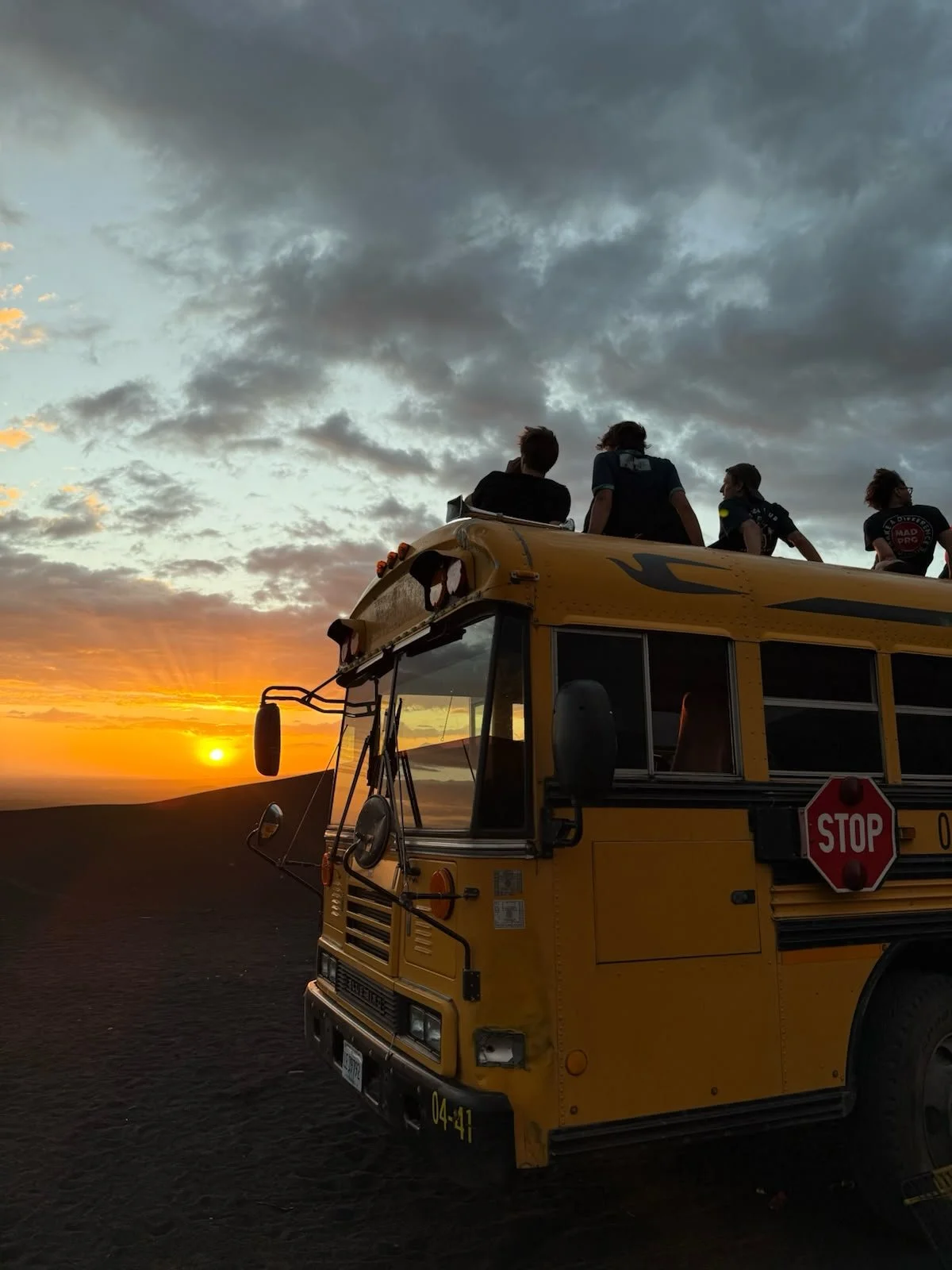 People sitting on top of a yellow school bus during a sunset, with a cloudy sky in the background.