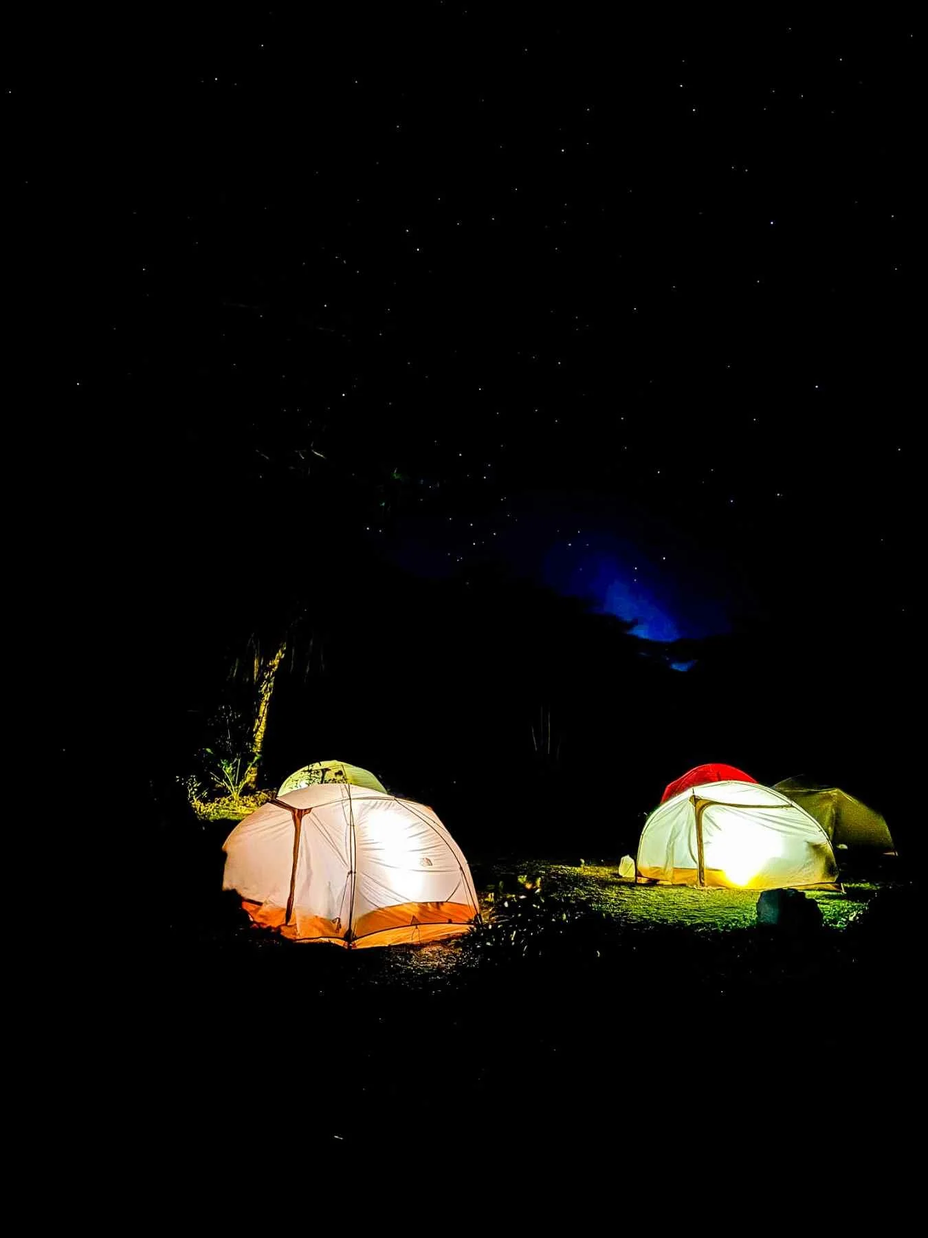 Nighttime scene of camping tents illuminated on the ground with a starry sky and a faint aurora in the background.