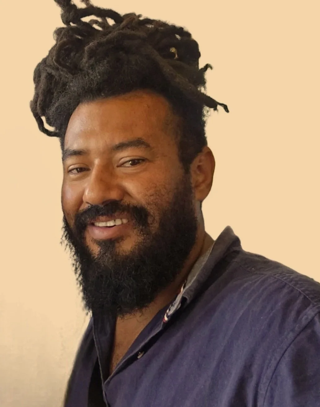 A man with a beard and dreadlocks smiling, wearing a dark button-up shirt, standing against a beige background.