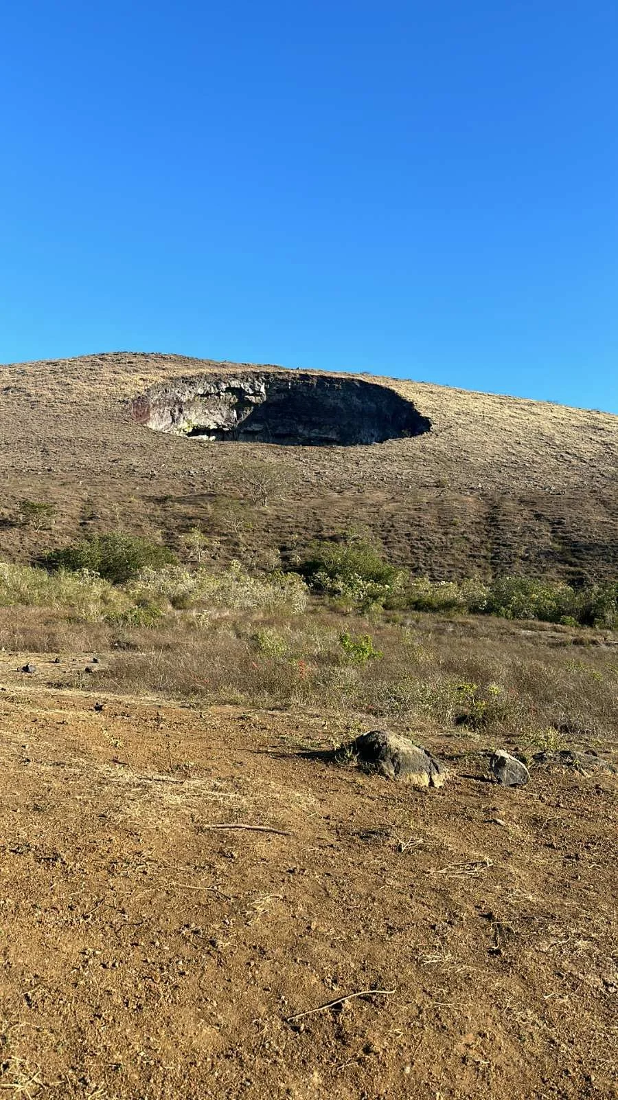 A dry, arid landscape with rocks and sparse vegetation, a hill or mountain in the background with a large dark crater or opening near its top, and a clear blue sky overhead.