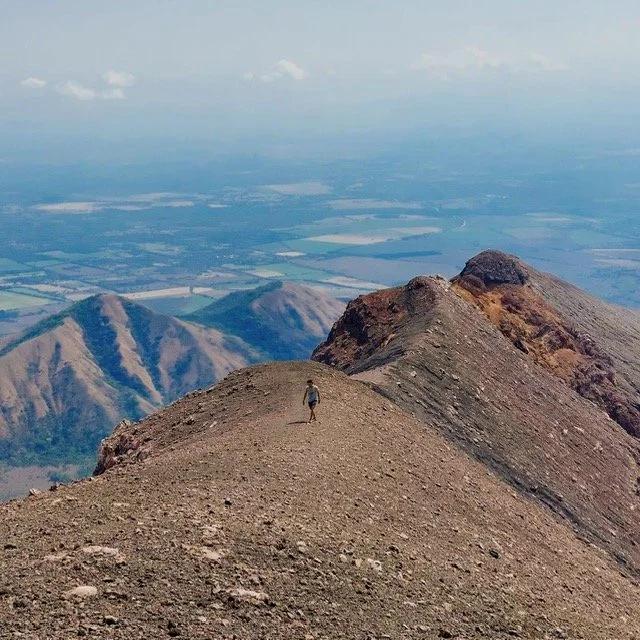 Hiker walking on a rugged volcano ridge of San Cristobal with a valley and distant volcanoes in the background.