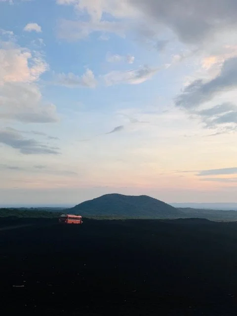 A landscape with dark foreground, a hill in the middle ground, and a sky with scattered clouds at sunset or sunrise.