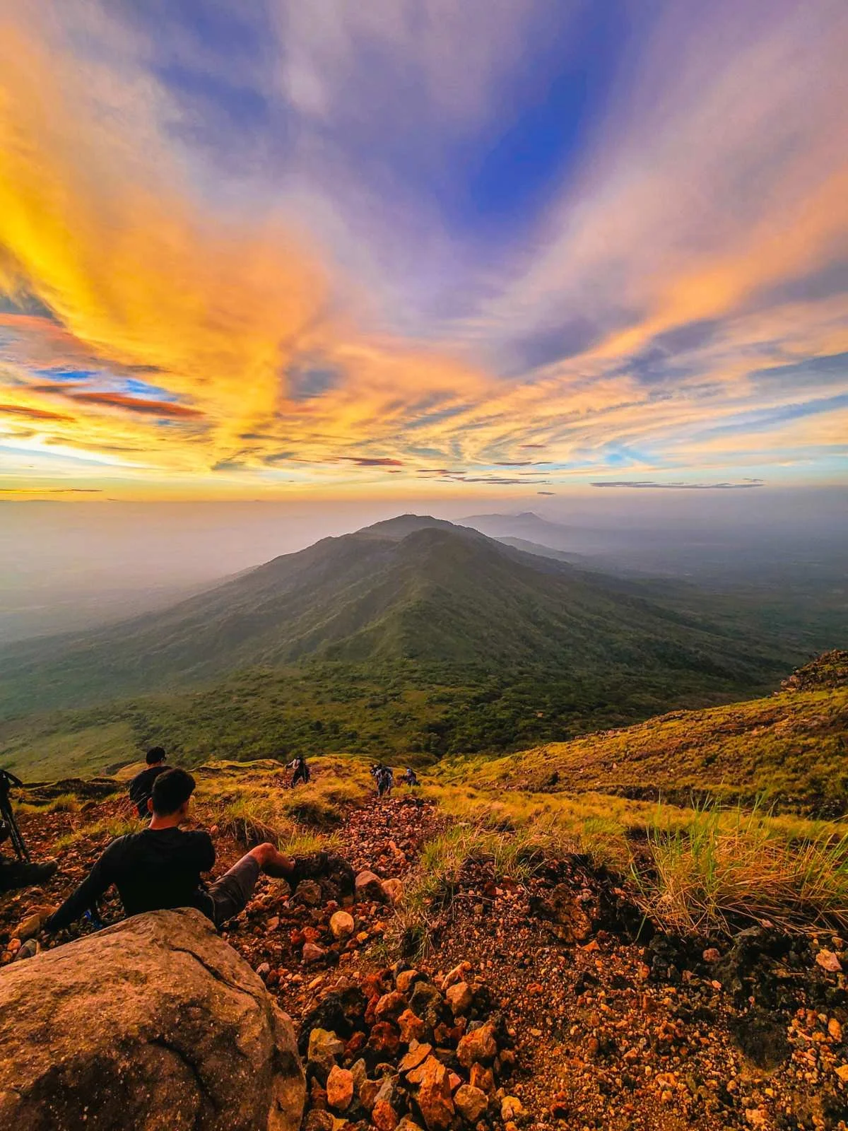 A scenic view of a mountain landscape during sunset with vibrant orange, pink, and purple clouds in the sky. In the foreground, people are sitting and walking on rocky terrain surrounded by green vegetation.
